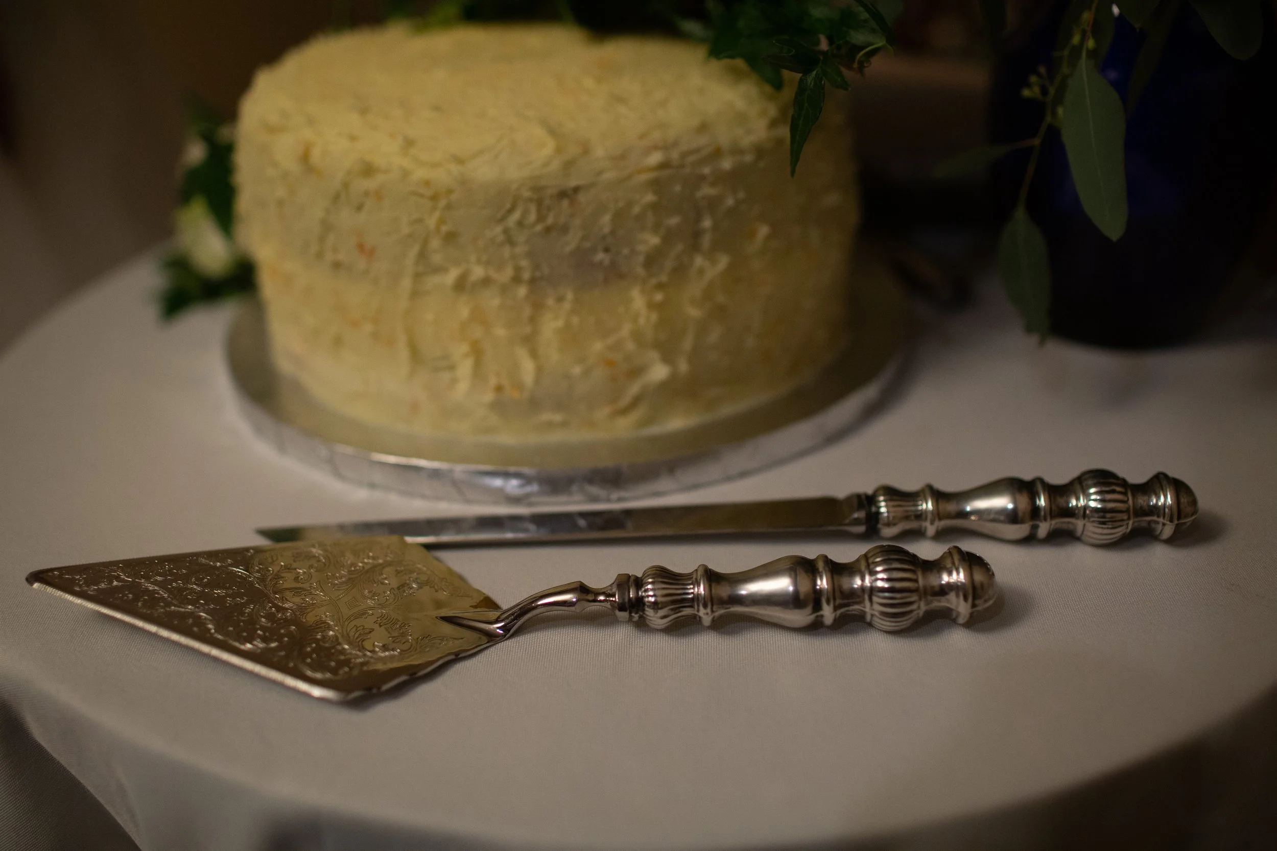 A close-up of a decorative cake server with an engraved handle, a knife with an ornate handle, and a cake on a plate, all on a white tablecloth.