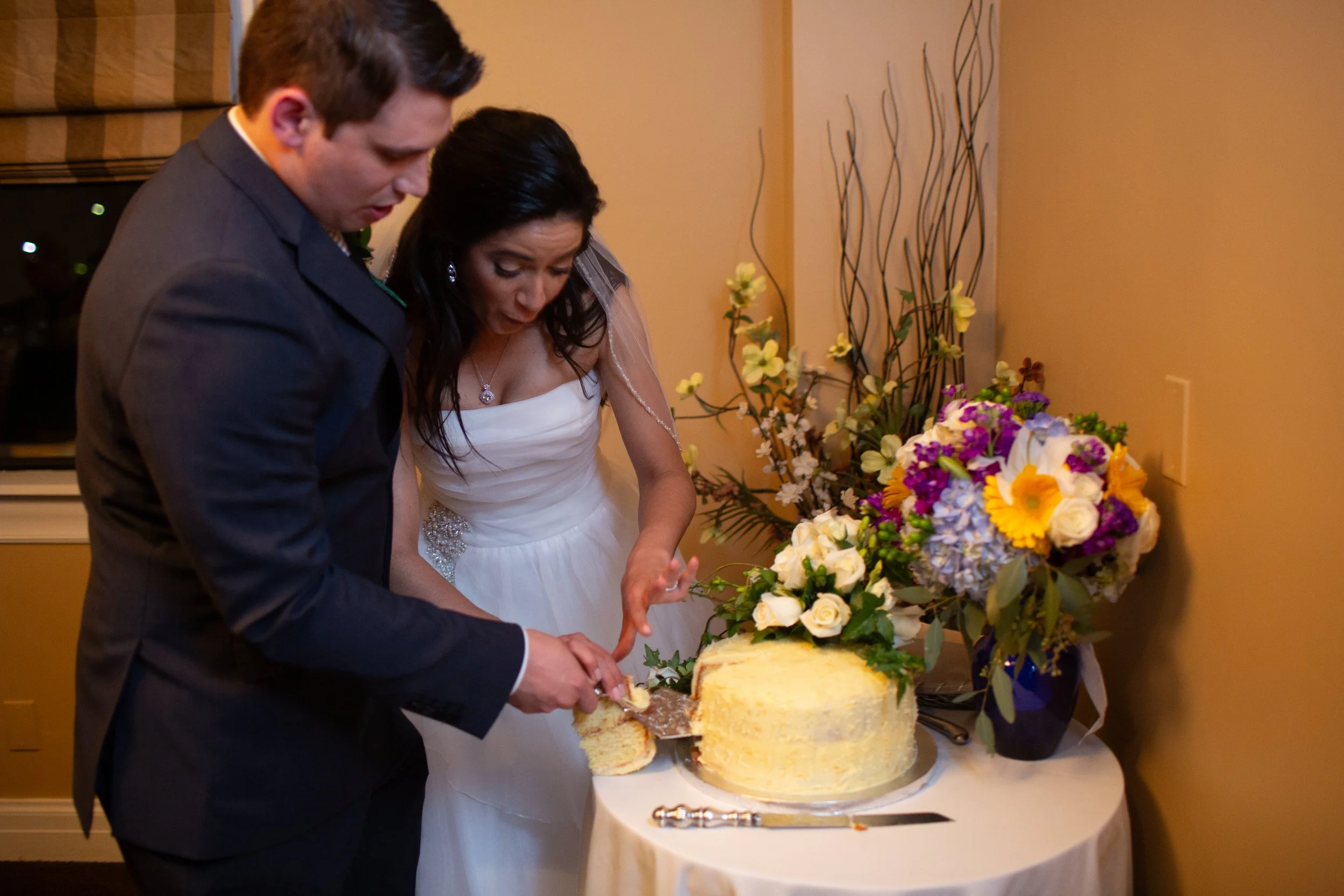 A bride and groom cutting a wedding cake together in a warmly lit room. The table is decorated with a large floral arrangement featuring yellow, white, purple, and pink flowers.