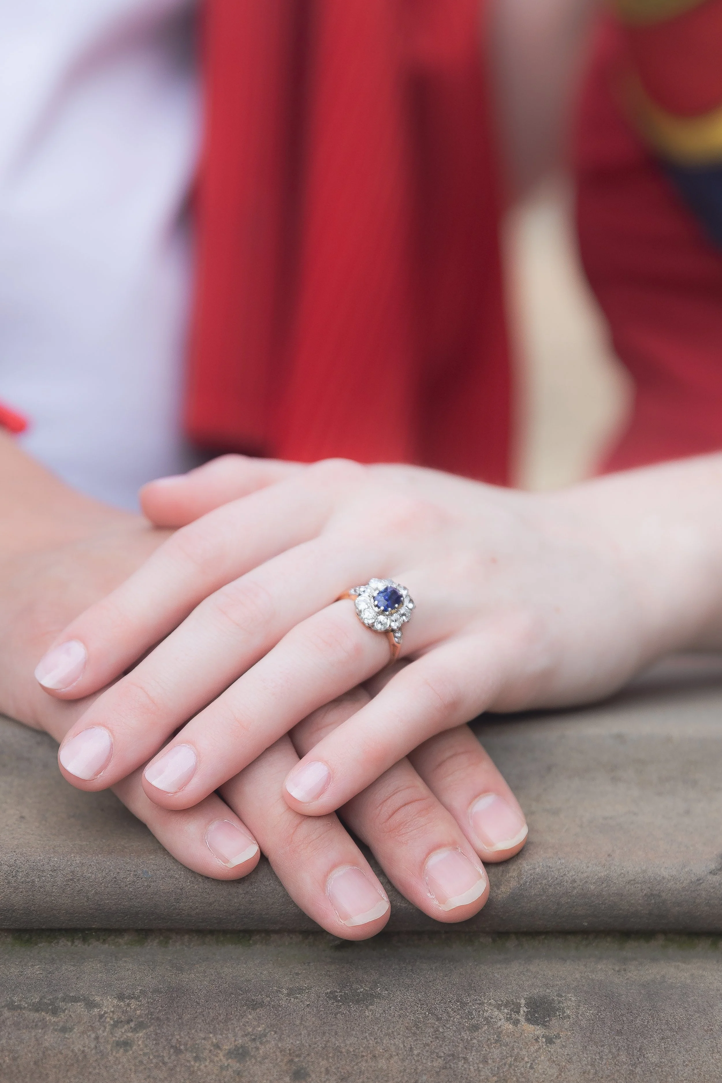 Close-up of two hands with one wearing a ring, resting on a brown surface, with a blurry background of a person in red and yellow clothing.