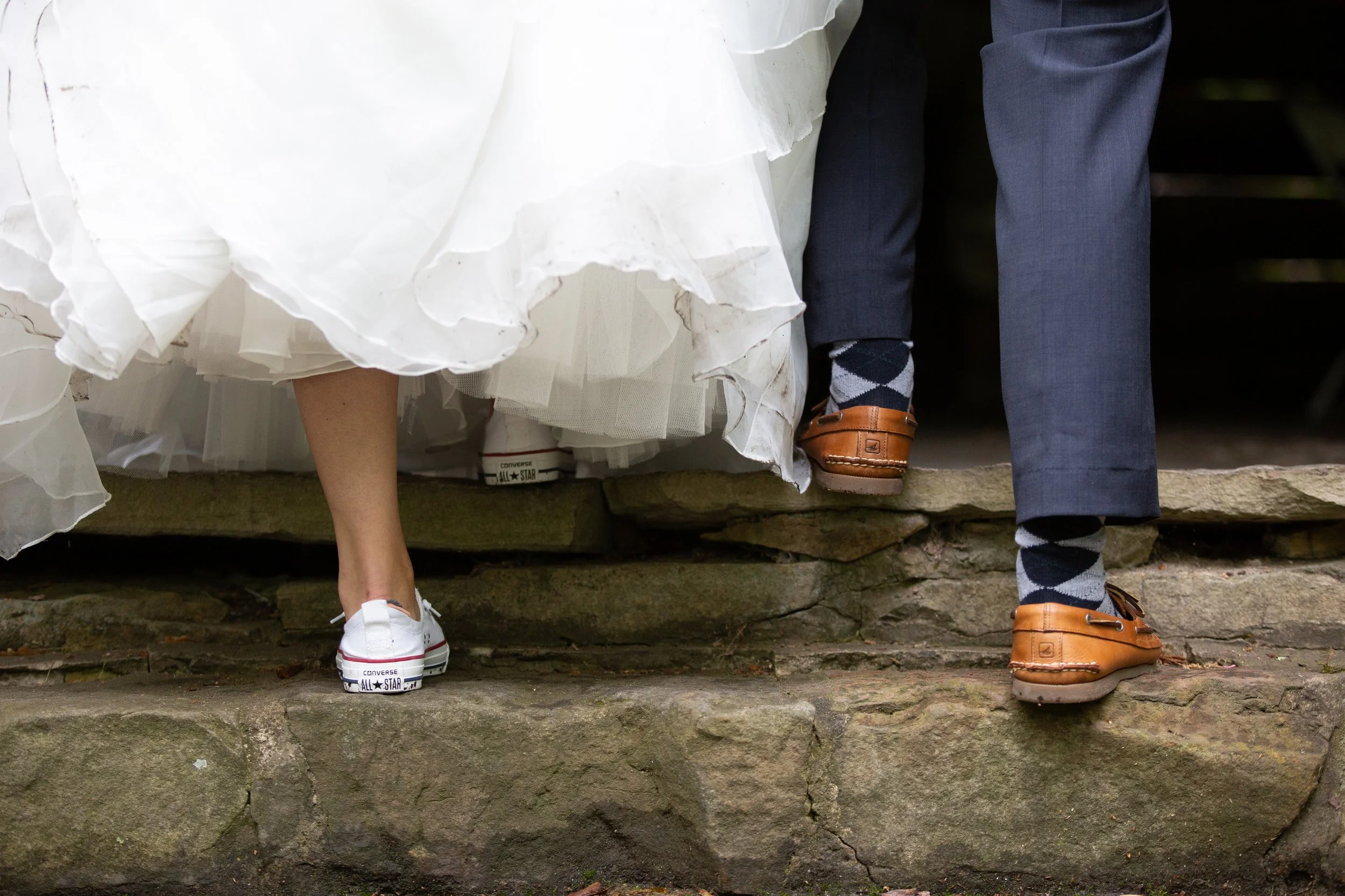 Close-up of the lower half of two people sitting on stone steps. The person on the left wears a white dress and white sneakers, while the person on the right wears gray pants, argyle socks, and brown loafers.