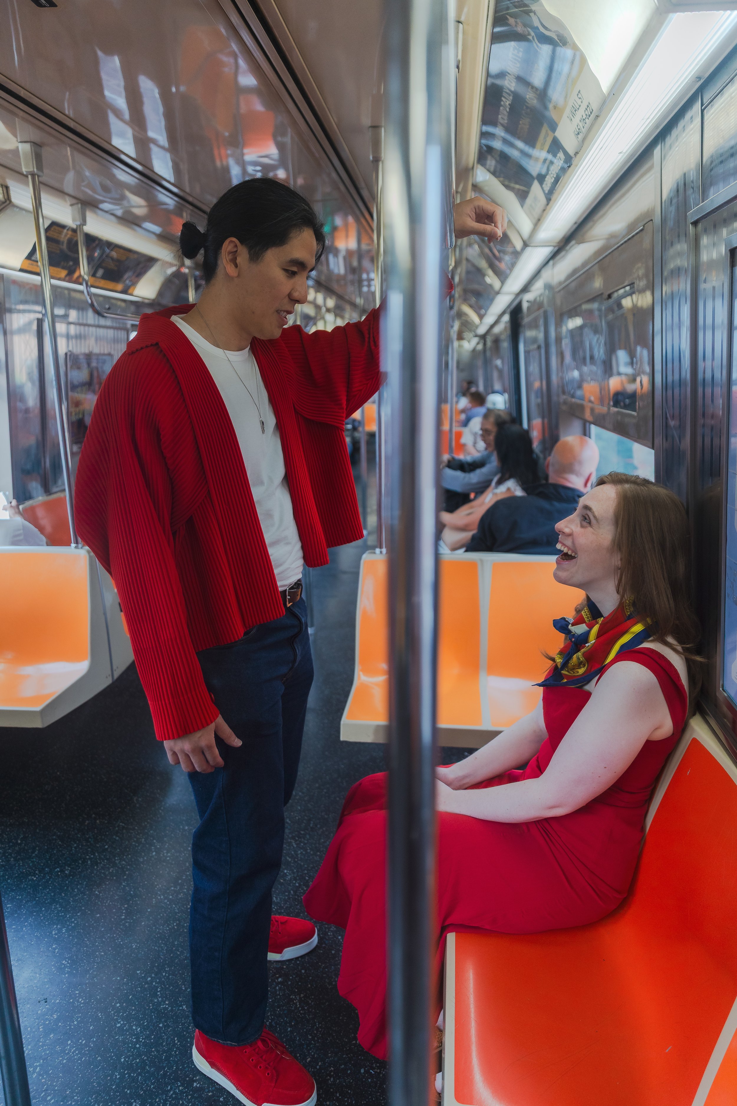 A man and a woman on a subway train engaging in conversation. The man is standing, leaning against a pole, while the woman is sitting and smiling up at him. The man is wearing a red jacket and red sneakers, and the woman is wearing a red dress with a