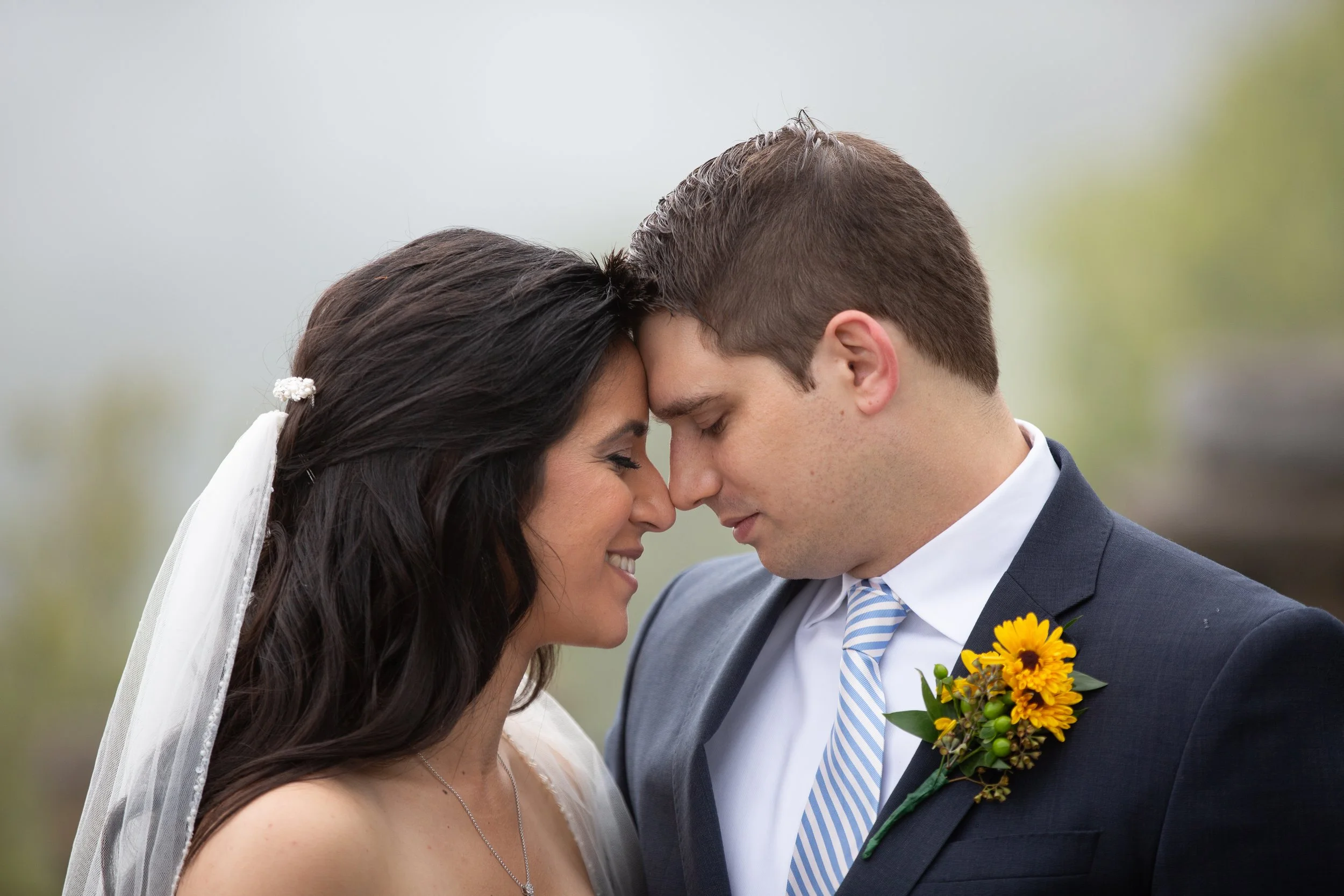 A bride and groom face each other with foreheads touching outdoors, both smiling softly, with the groom wearing a dark suit and striped tie, and the bride in a white dress and veil, holding a bouquet of yellow flowers.