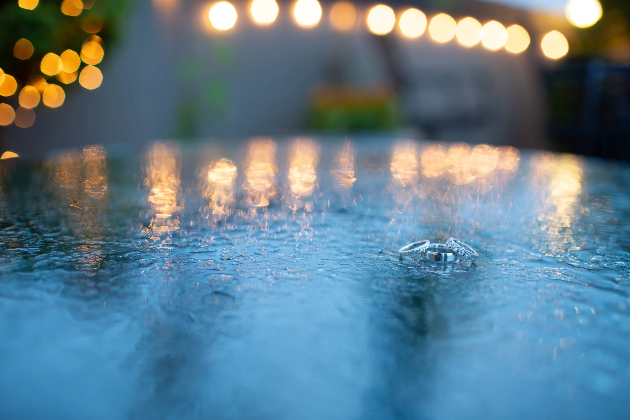 A close-up of wedding rings resting on a wet surface with blurred warm bokeh lights in the background.