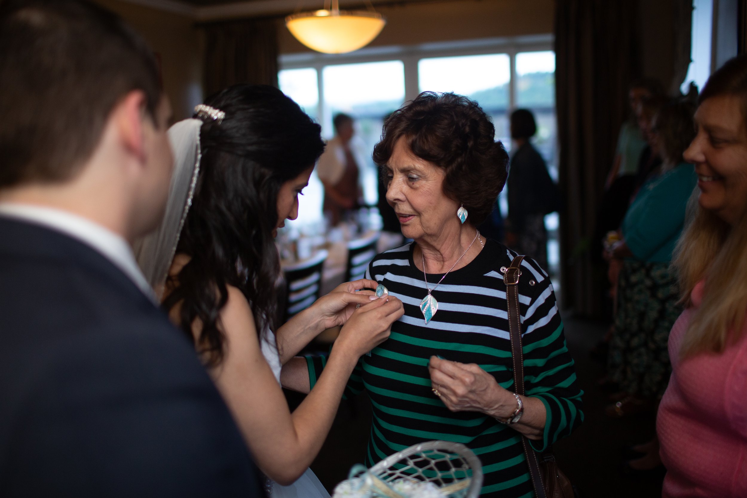 A woman with short hair and a striped top is receiving jewelry from a bride during a wedding celebration. The bride is wearing a wedding dress and veil, and the woman is smiling warmly at the jewelry being presented to her. Other guests are visible i