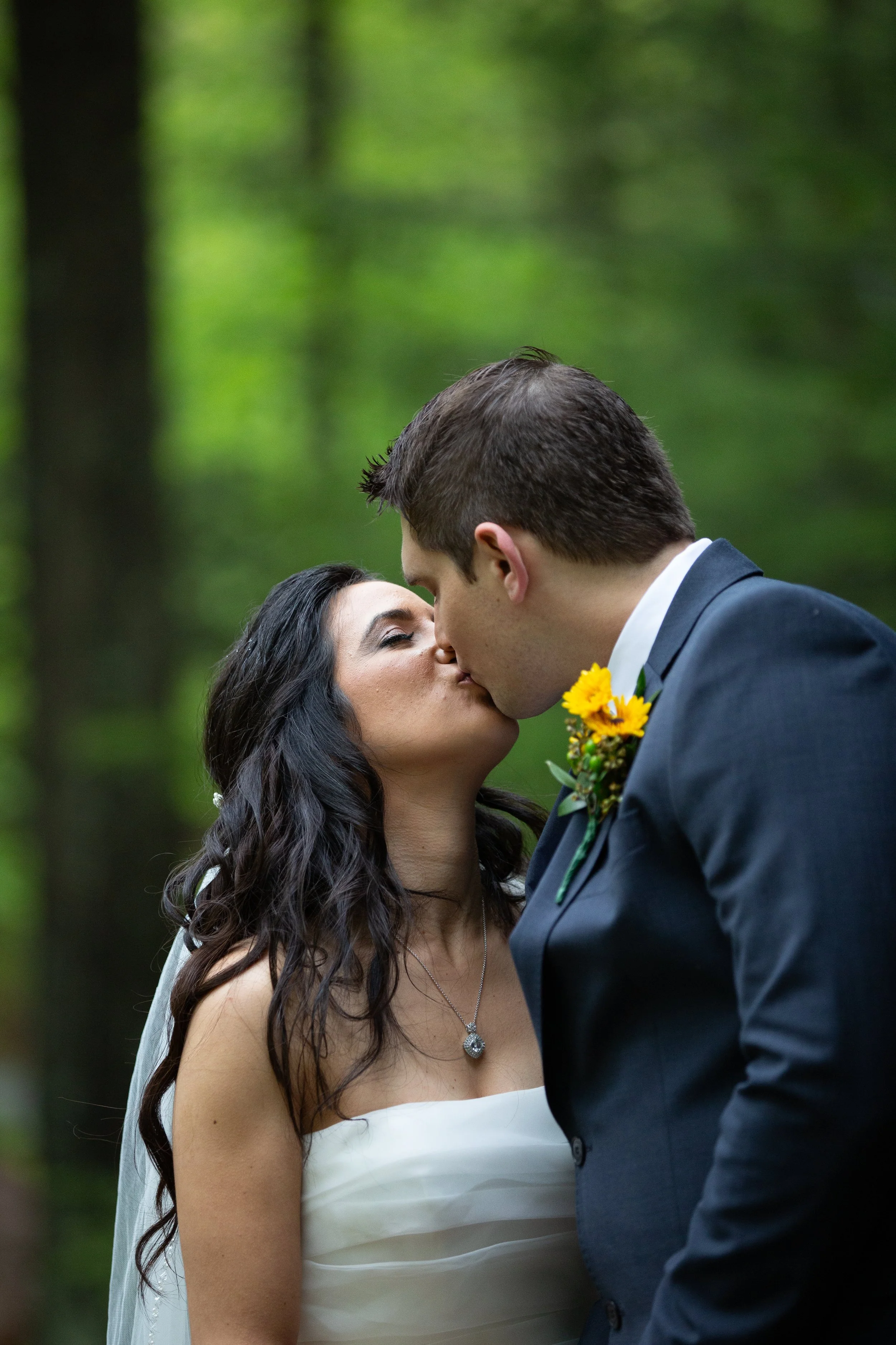 A bride and groom are about to kiss in a forested outdoor setting, with the bride wearing a strapless white dress and a silver necklace, and the groom in a dark suit with a flower boutonniere.
