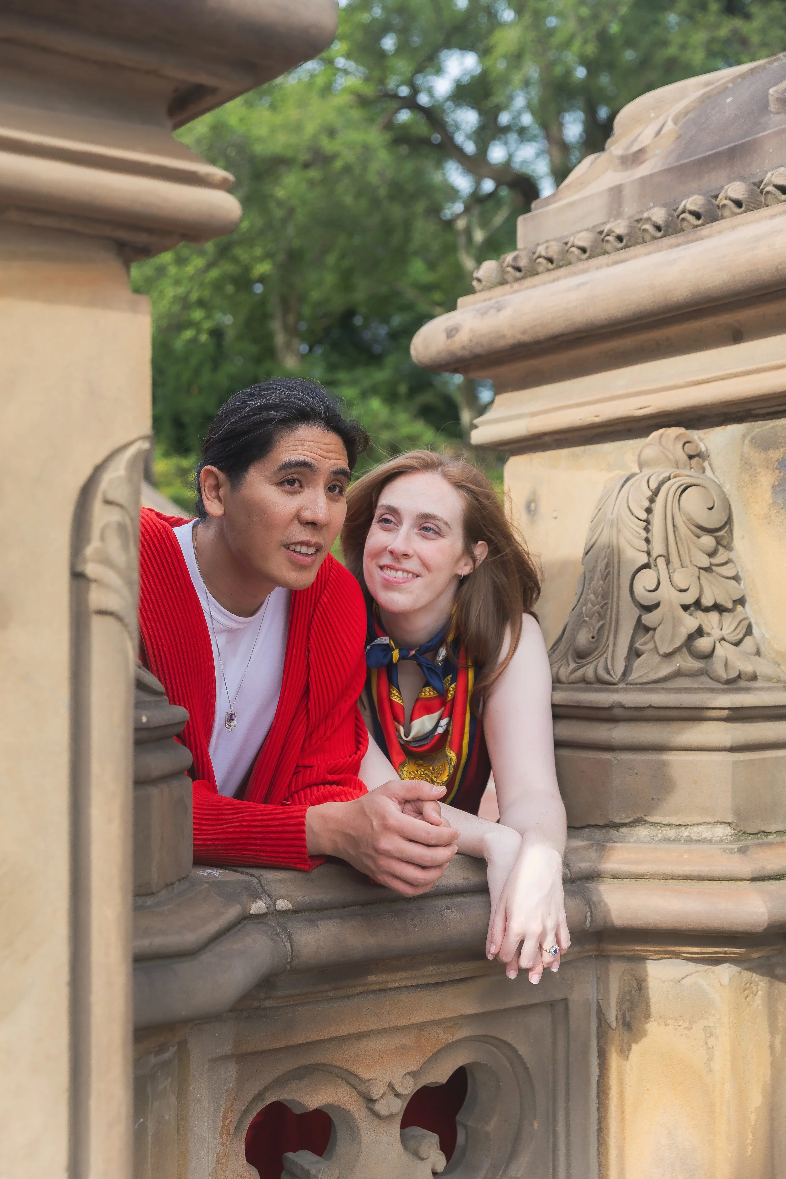 Two people leaning on a stone balcony, both smiling, outdoors with green trees in the background. The person on the left is a man with dark hair wearing a red jacket, and the person on the right is a woman with light brown hair wearing a colorful sca