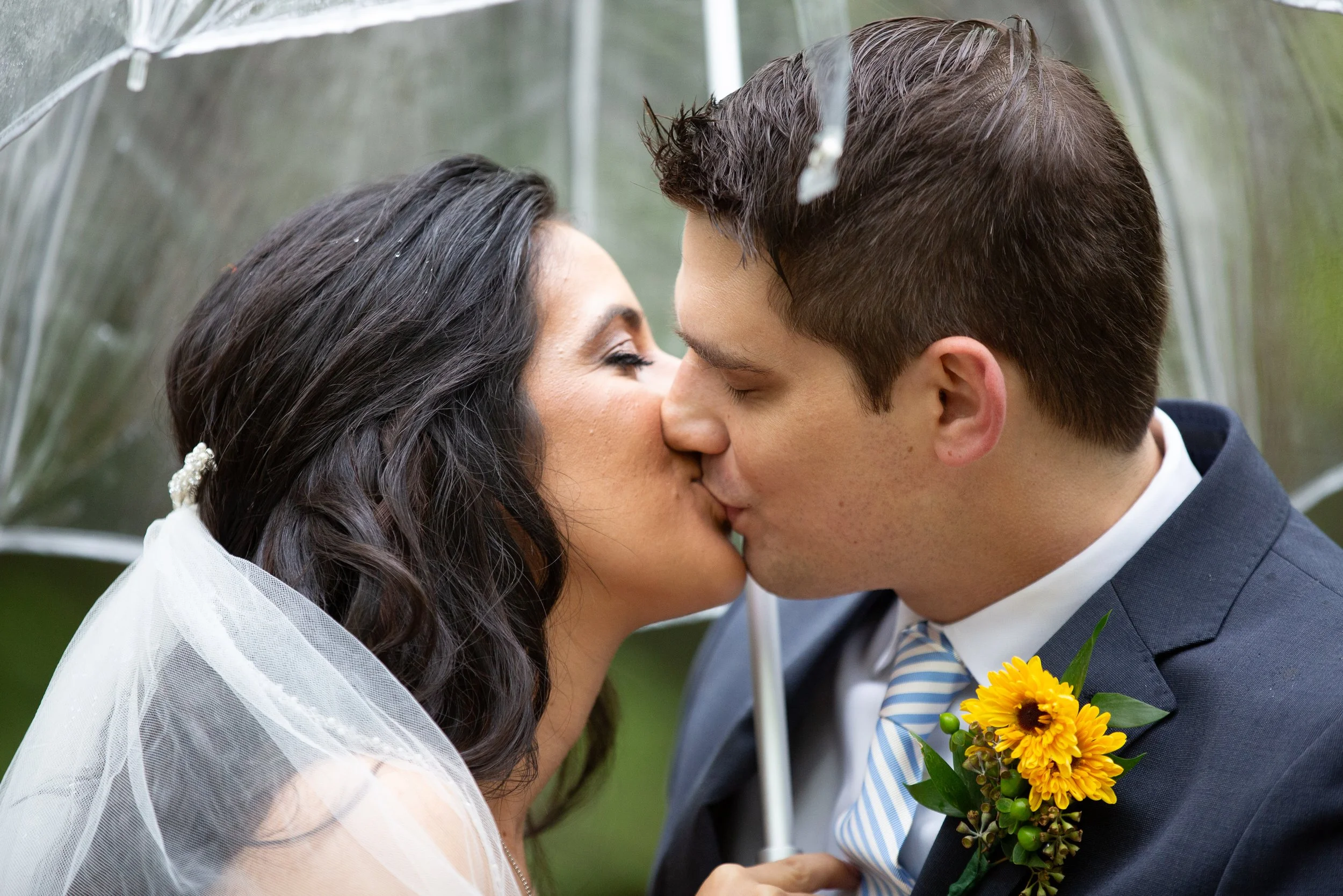 A bride and groom are sharing a kiss outdoors. The bride has long dark hair with a veil, and the groom has short brown hair and is wearing a suit with a striped tie, holding a bouquet of yellow flowers.