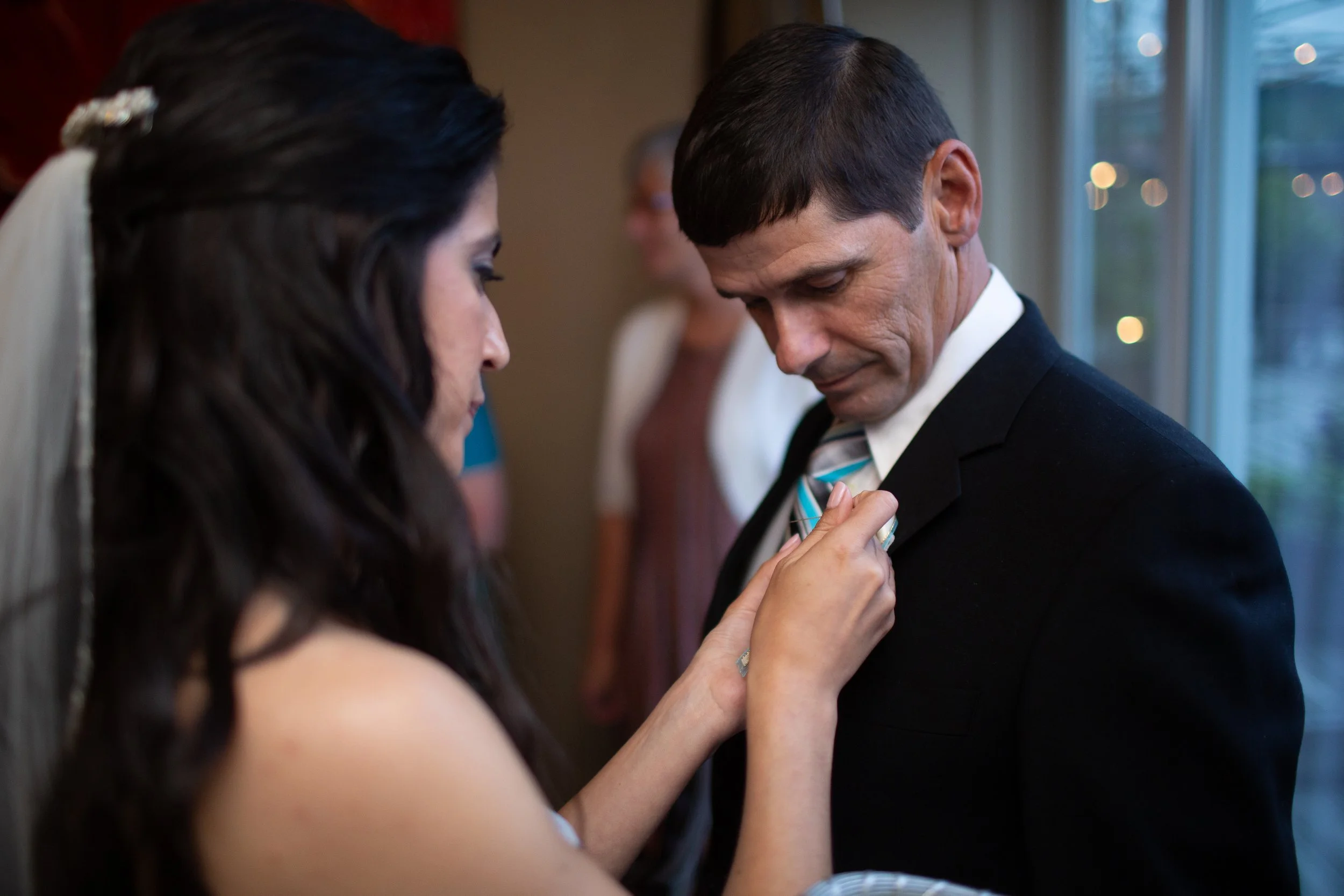 A woman pinning a boutonniere on a groom at a wedding, with other guests in the background.
