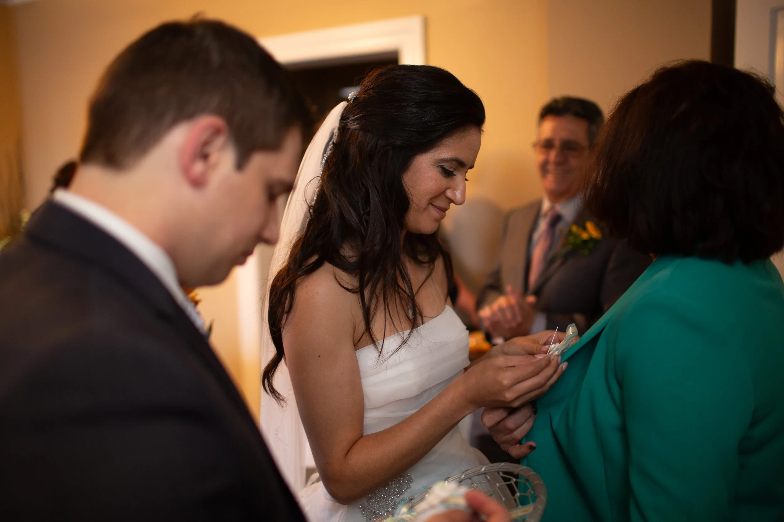 A bride in a white wedding dress with long dark hair is smiling and holding hands with a woman in a teal blazer during a wedding ceremony. A man in a suit is in the foreground, and a man in glasses and a suit is smiling in the background.