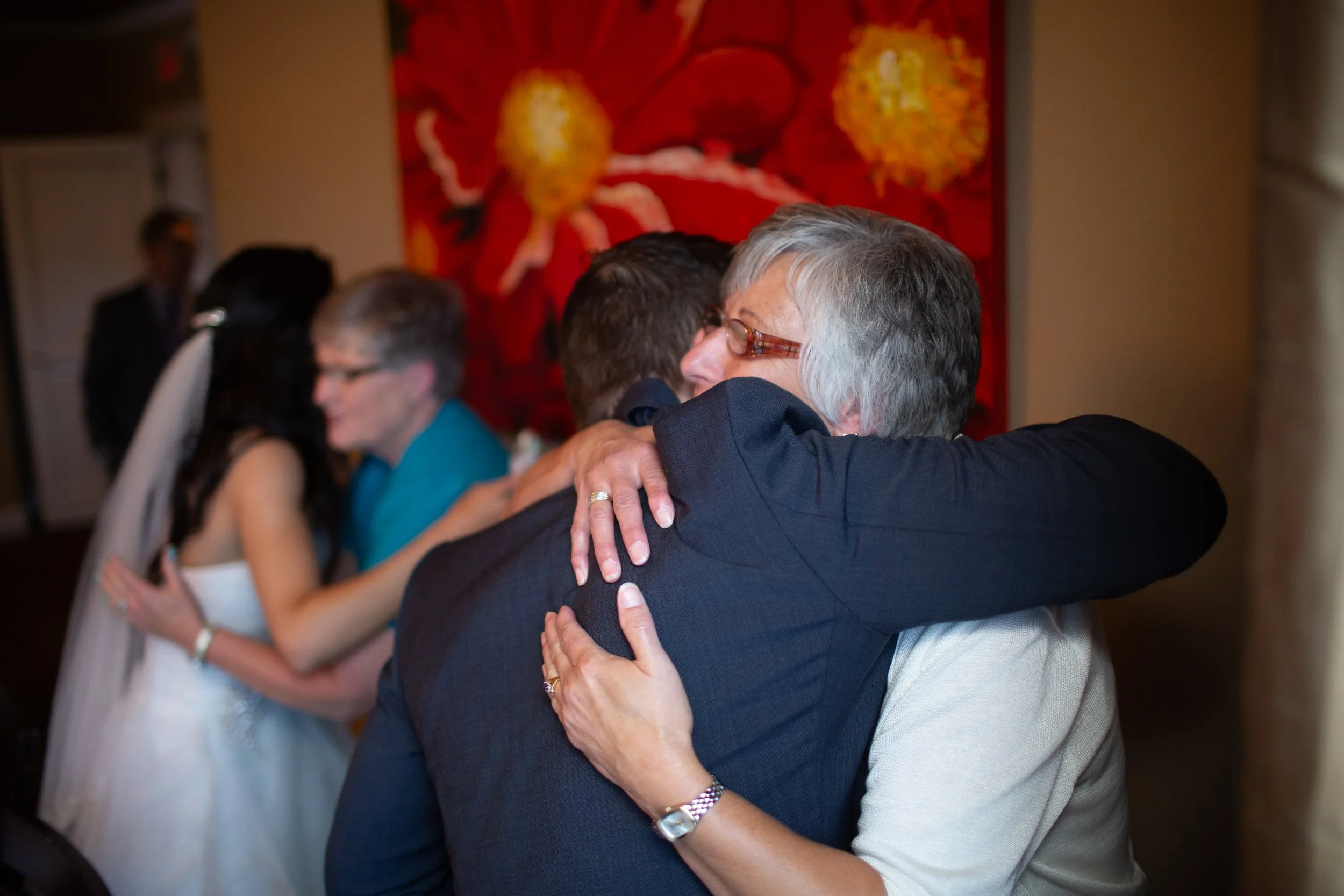 People hugging each other in a close embrace at an indoor event, with a colorful floral artwork in the background.