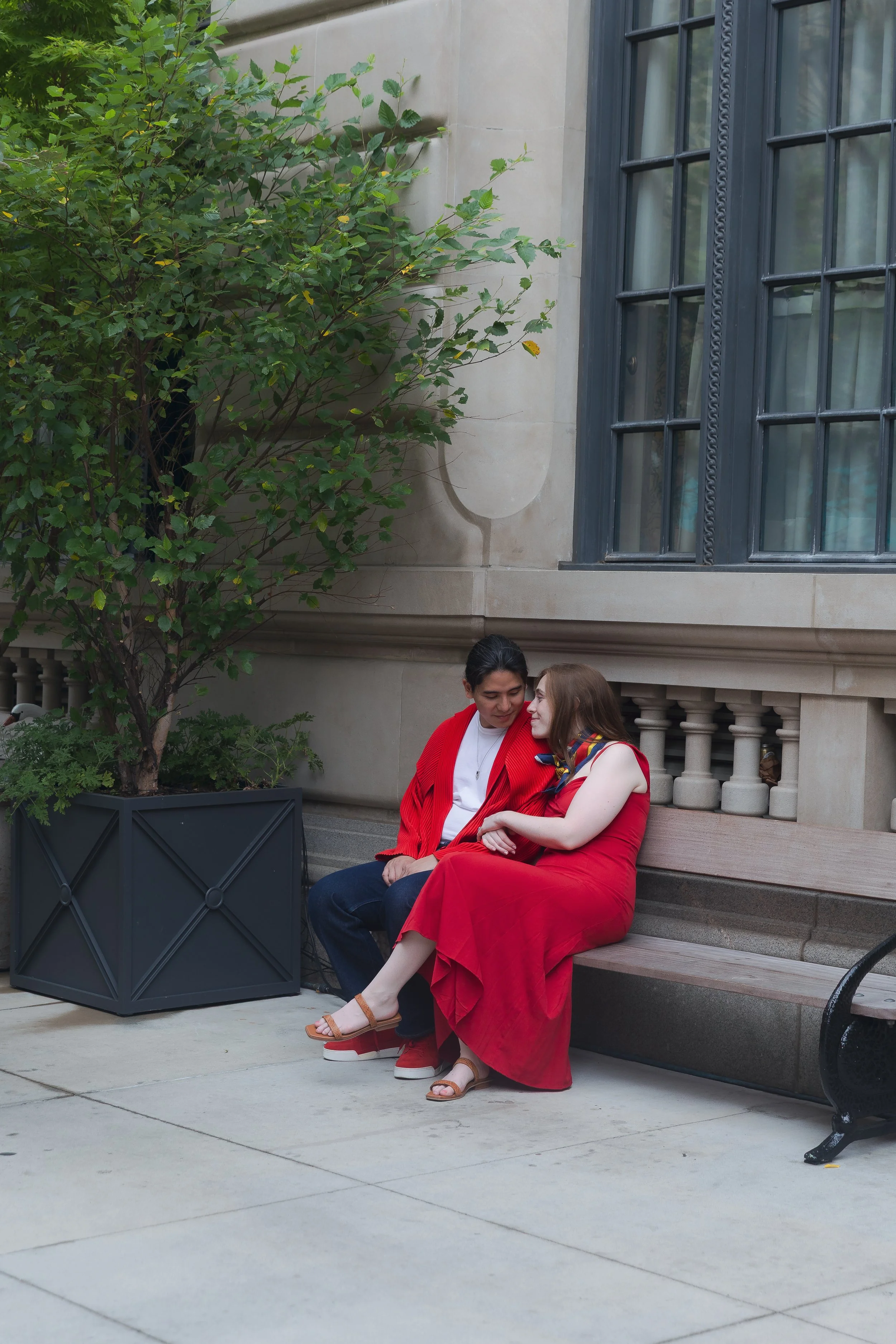 A young couple sitting on a wooden bench outside a building with large windows and stone walls, under a leafy tree, dressed in red and smiling at each other.