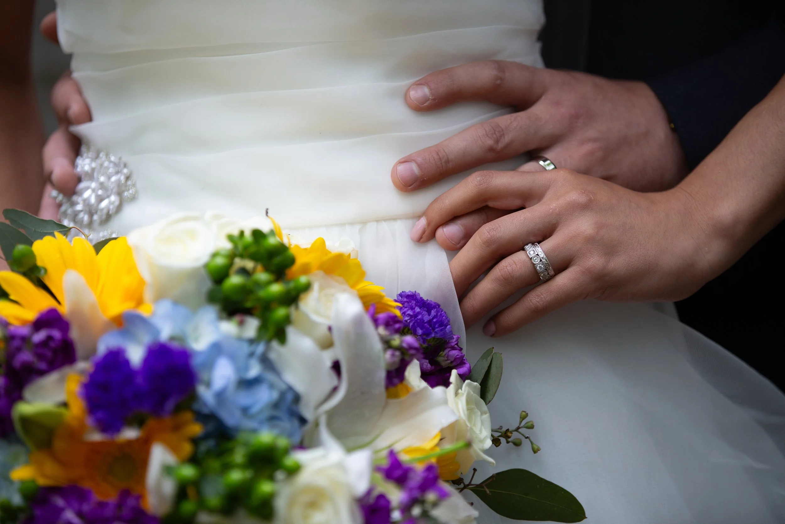 A close-up of a couple's hands on a white wedding dress, with wedding rings visible, surrounded by colorful flowers.