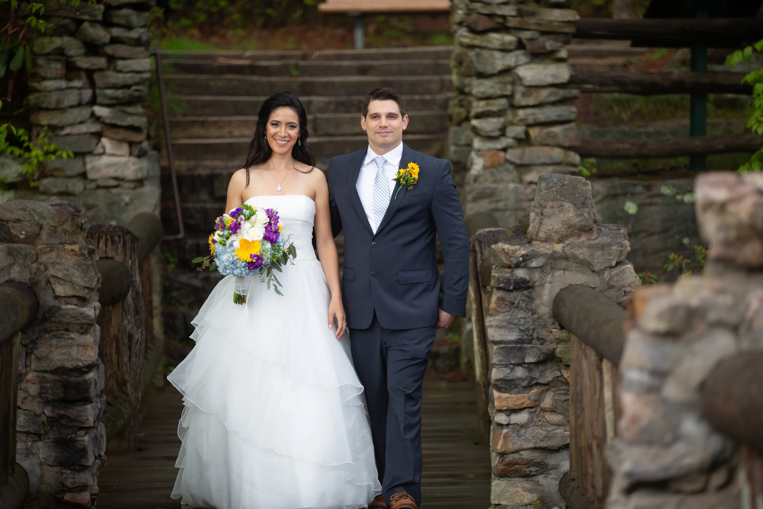 A bride and groom standing on a stone bridge outdoors, smiling at the camera. The bride is holding a colorful bouquet, and the groom has a yellow flower boutonniere.