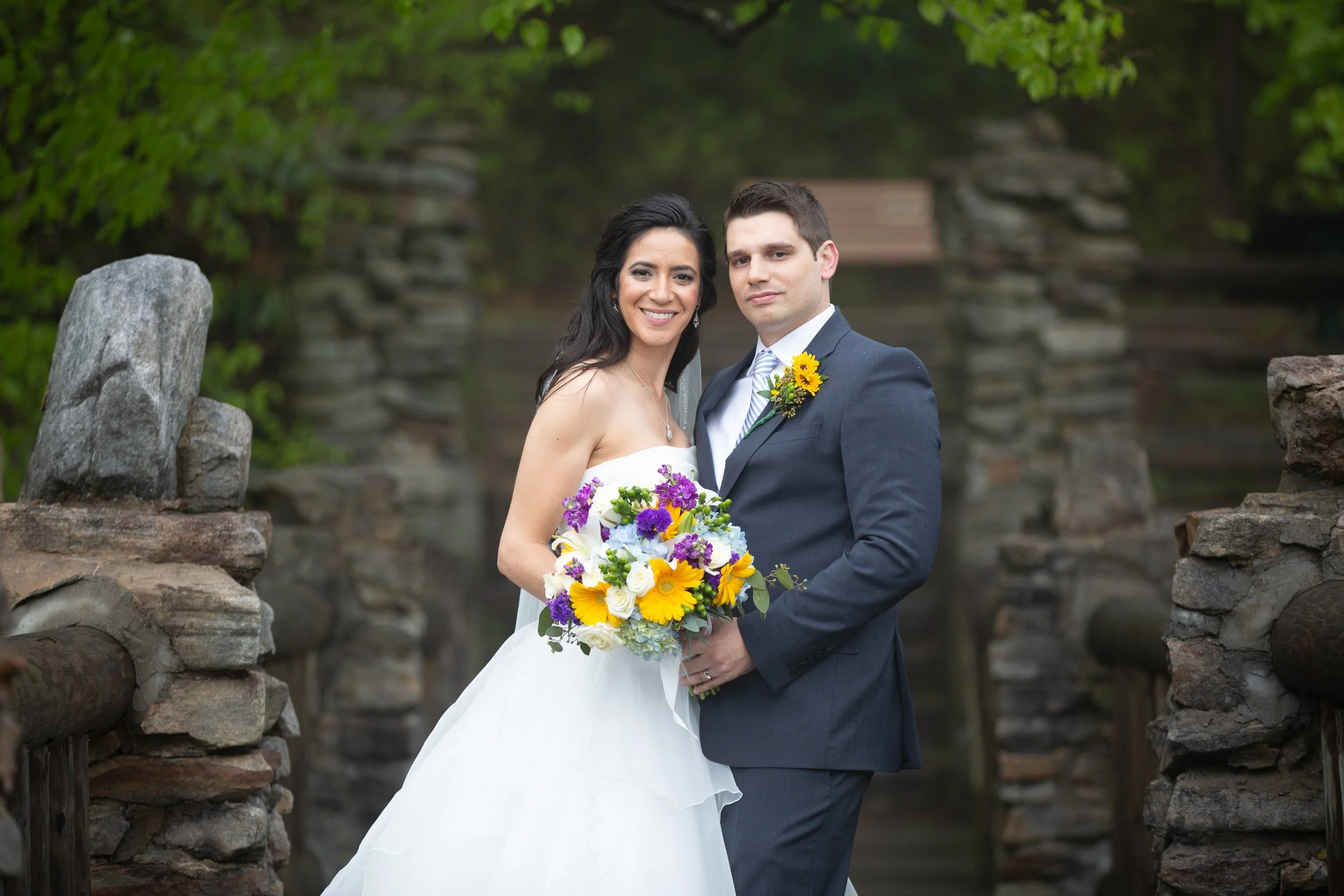 A bride and groom standing close together outdoors, smiling, with greenery and a stone structure in the background. The bride is holding a colorful bouquet and is wearing a strapless white wedding dress. The groom is dressed in a dark suit, white shi