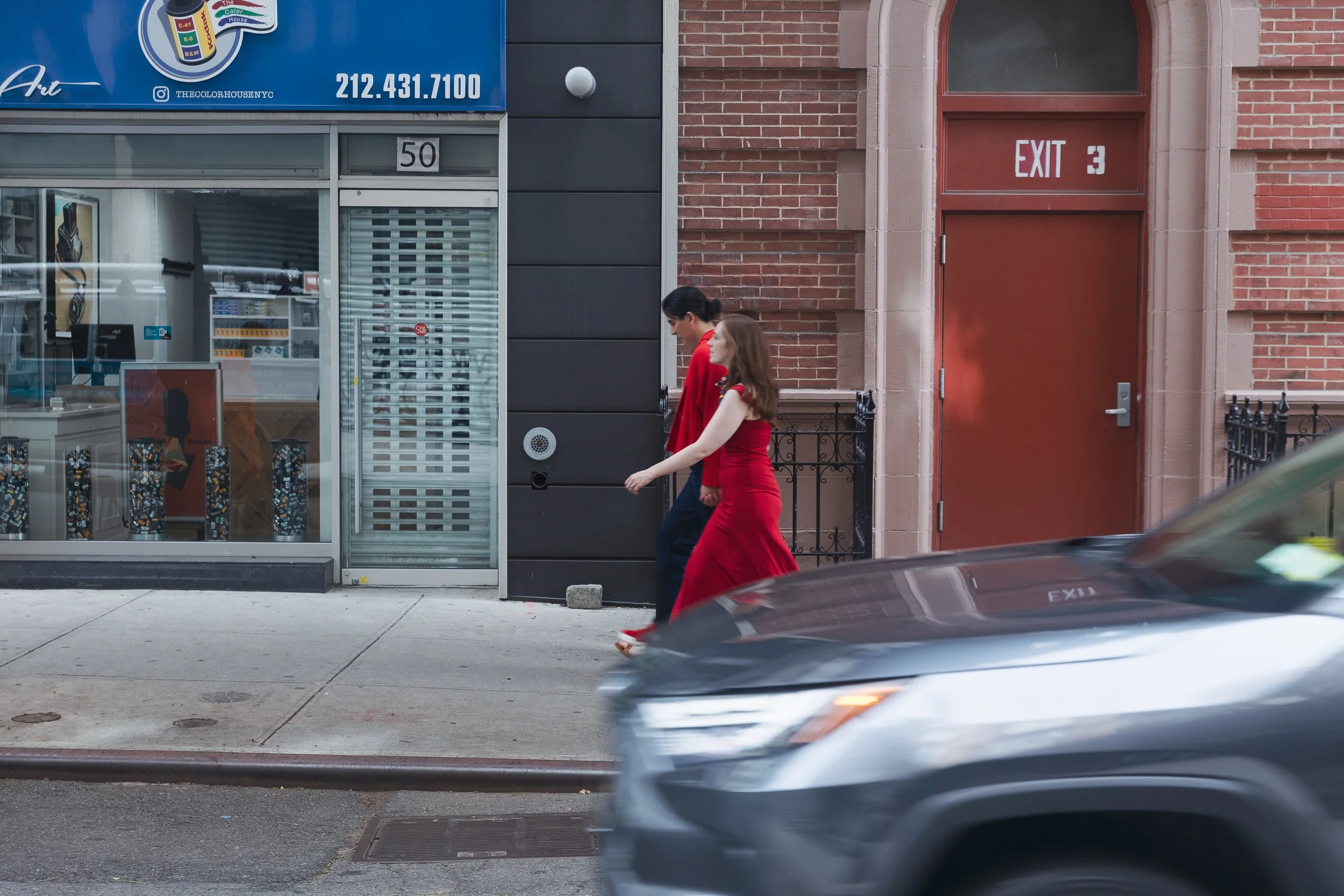 A woman in a red dress and a man in a dark suit walk past storefronts on a city sidewalk, with a blurry car passing in the foreground.