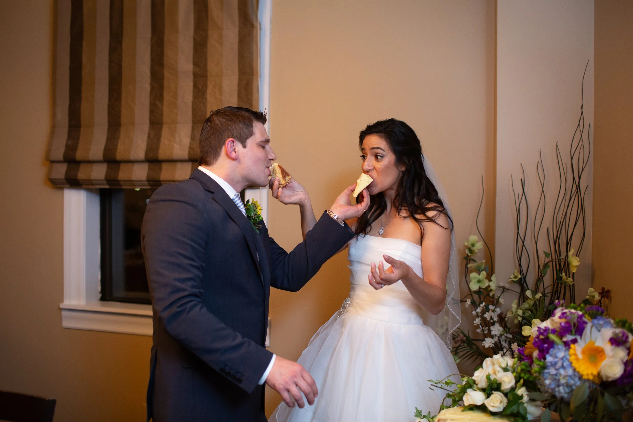 A bride and groom sharing a cake during their wedding reception, with flowers in the background.