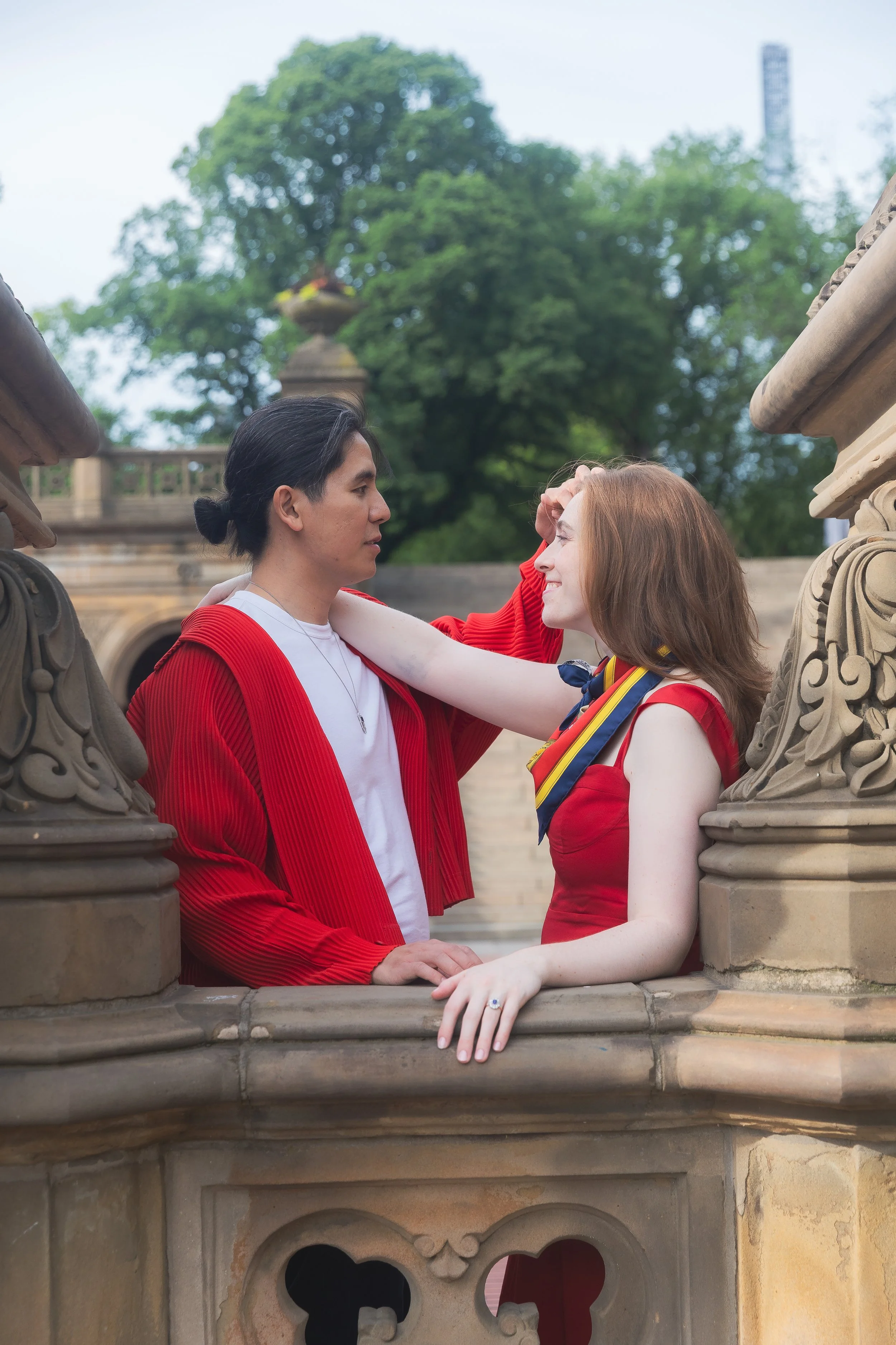 A man and woman face each other on a stone balcony, with ornate stone balustrades, surrounded by trees with a city skyline in the background. The man has black hair pulled back, wearing a white shirt and red jacket. The woman has red hair, wearing a 