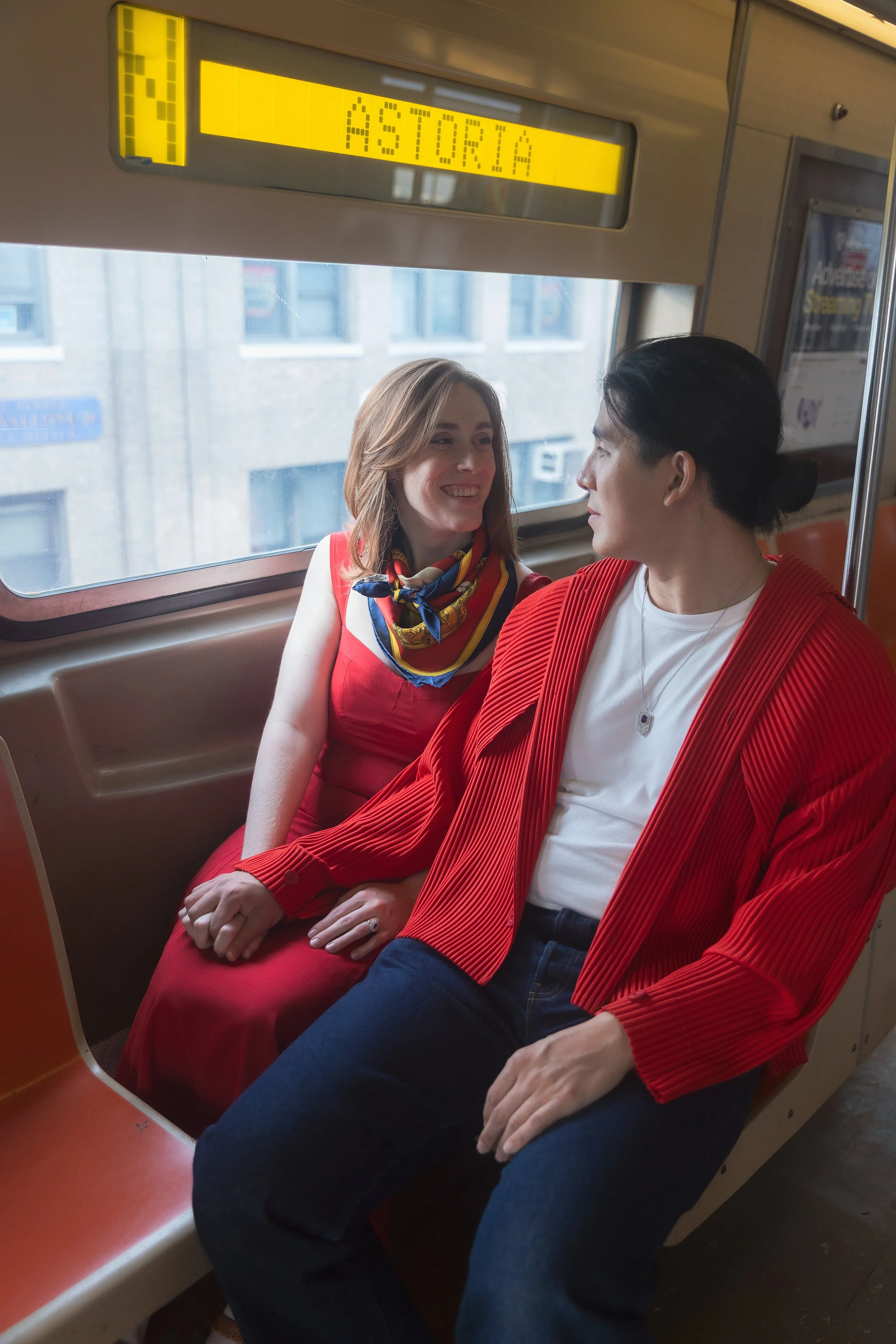 A couple sitting close together on a bus, facing each other and smiling. One is wearing a red dress with a yellow scarf, and the other is in a white T-shirt and a red jacket. The background shows windows and a cityscape.