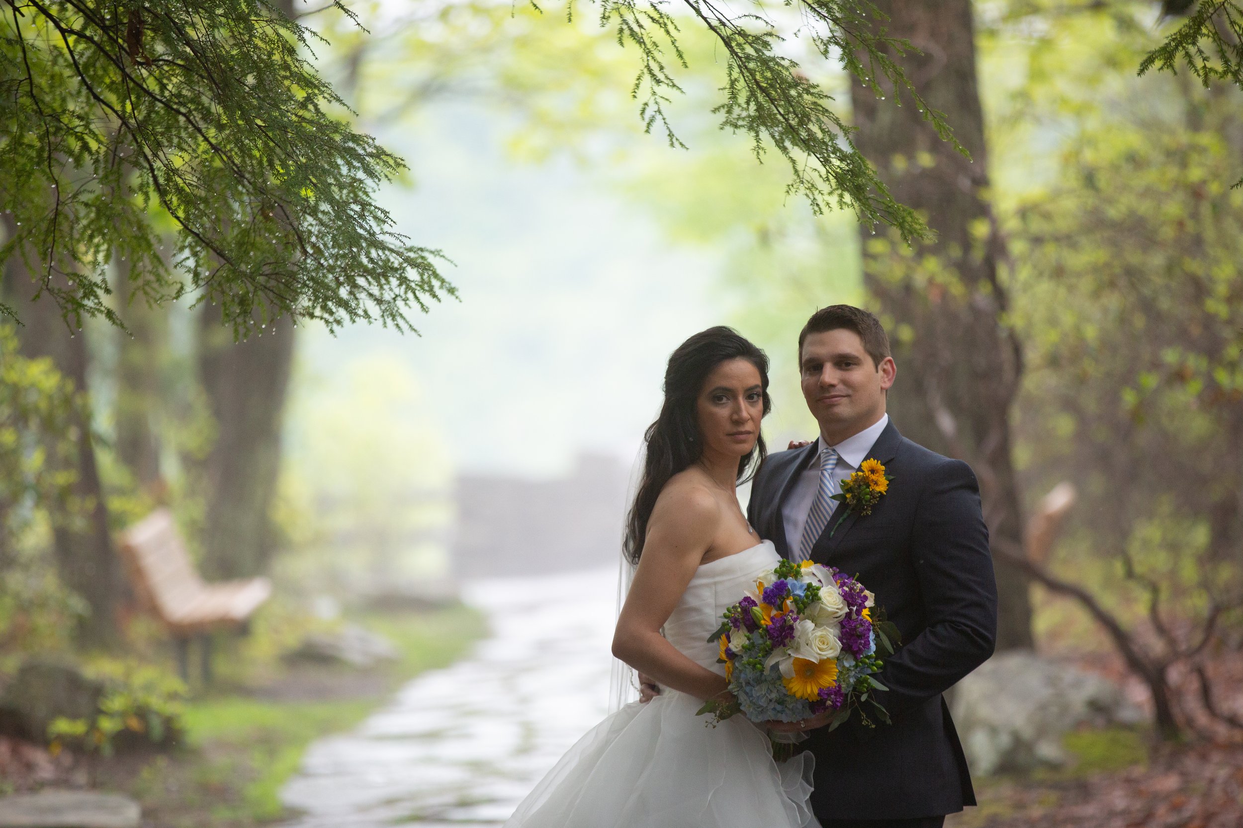 A bride and groom standing together outdoors in a lush, green forest with trees and a small water feature in the background, the bride holding a colorful bouquet and the groom wearing a suit with a yellow flower boutonniere.