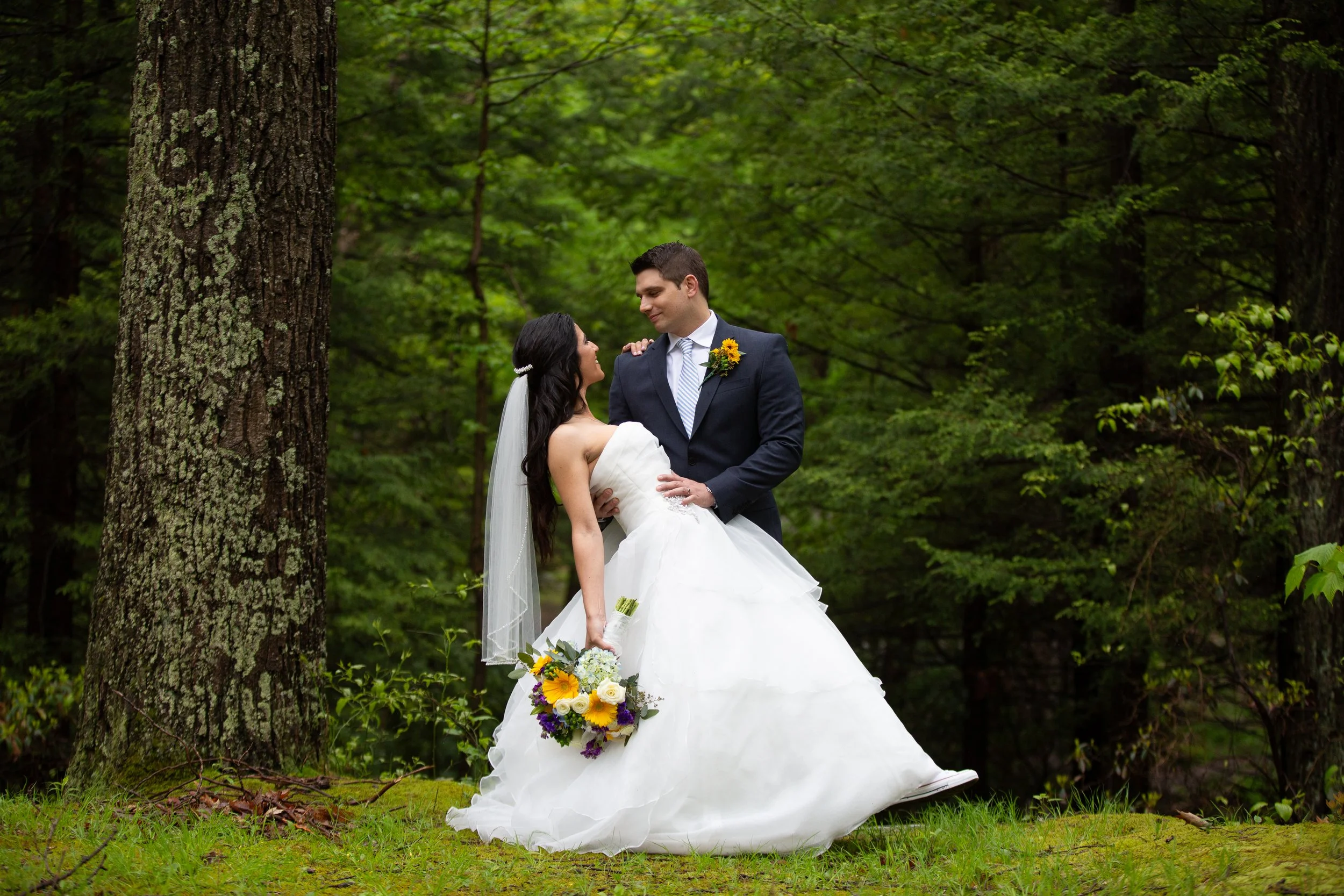 A bride and groom are outdoors in a lush green forest. The bride is in a white wedding dress and holding a bouquet. The groom is in a dark suit with a striped tie and a sunflower boutonniere. They are smiling and looking at each other lovingly.