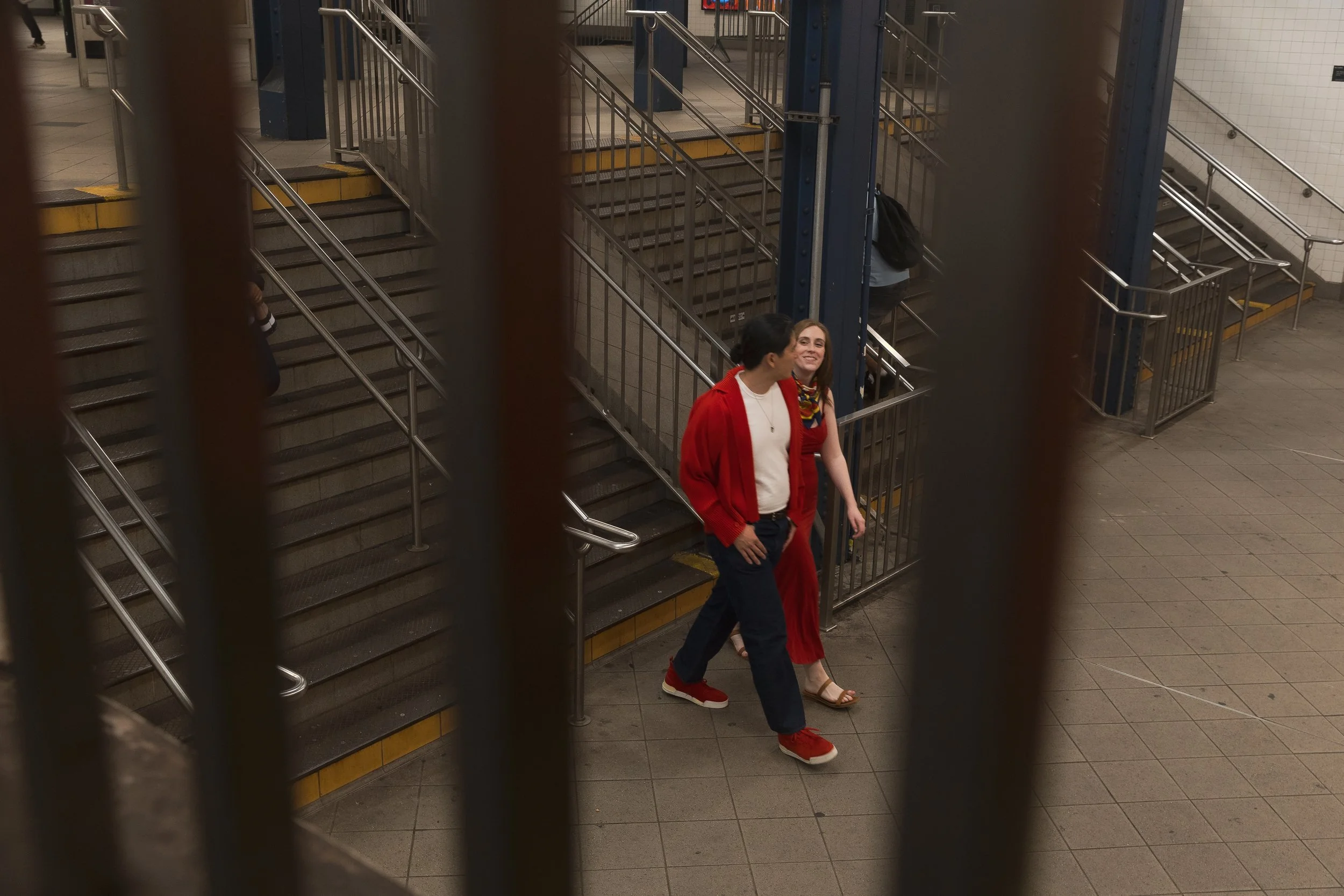 Two people walking up steps in a subway station, seen through metal bars. One woman wears a red cardigan and dark pants, and the other woman wears a colorful scarf, beige sandals, and a sleeveless top.