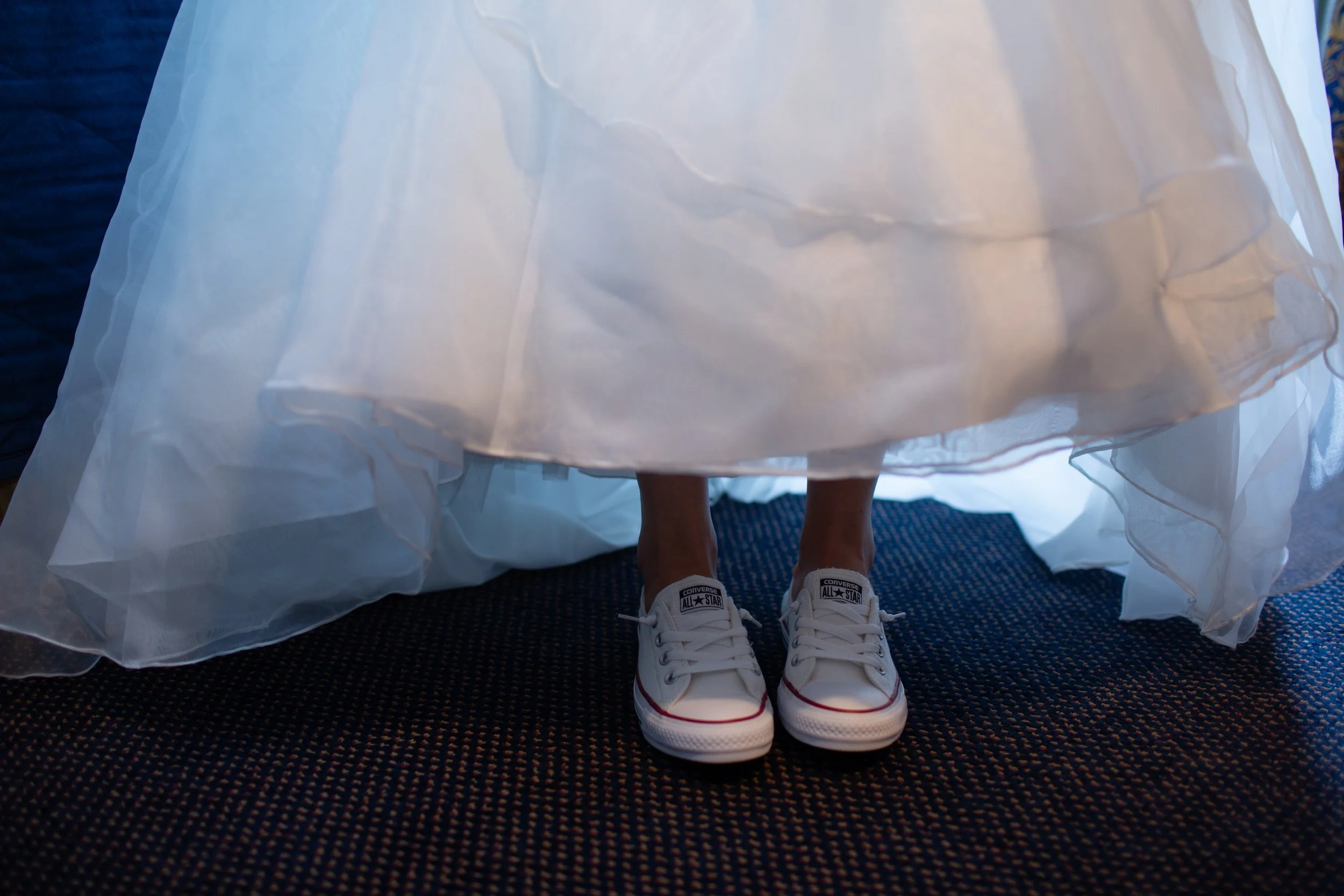 Person standing on a dark carpeted floor, wearing white Converse sneakers and a white dress, visible through the sheer fabric of the dress.