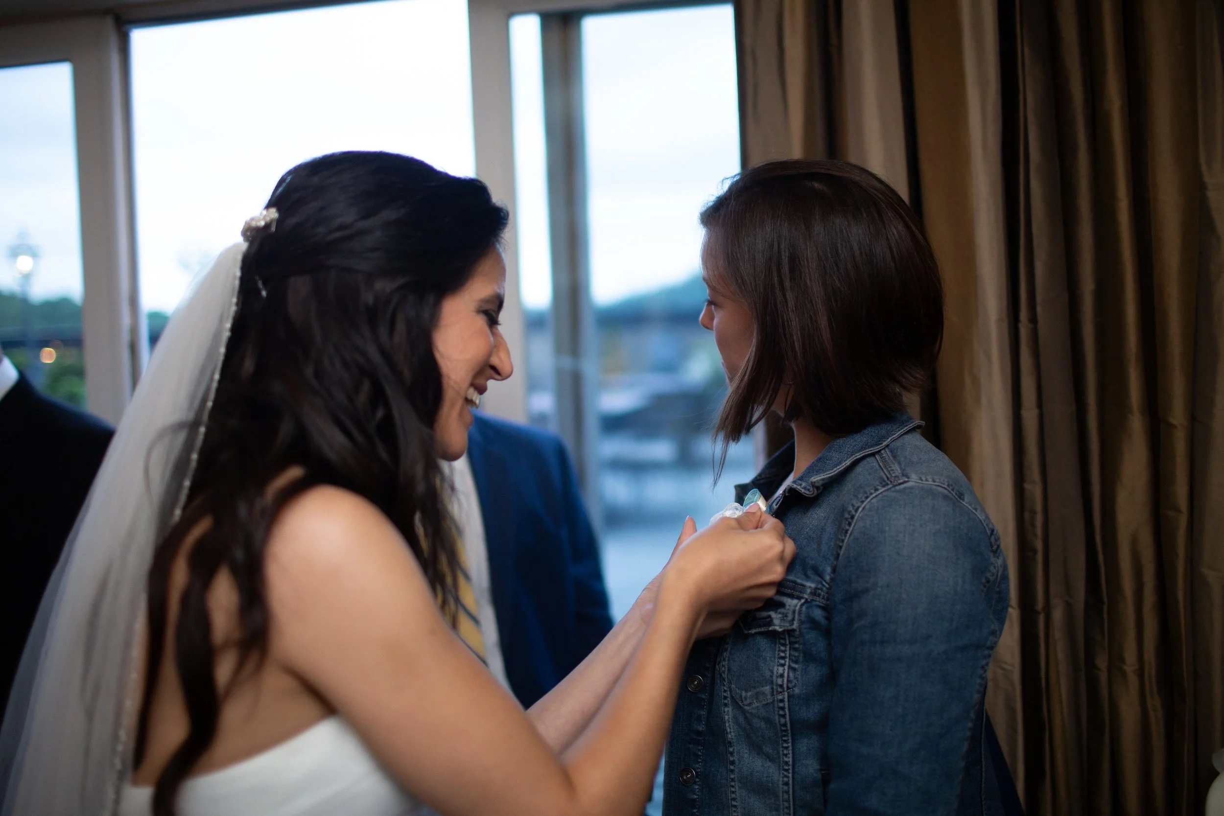 A woman with long dark hair, wearing a white dress and veil, is smiling and leaning forward as another woman with short brown hair, wearing a denim jacket, helps her pin a badge or flower to her clothing inside a room with large windows and brown cur