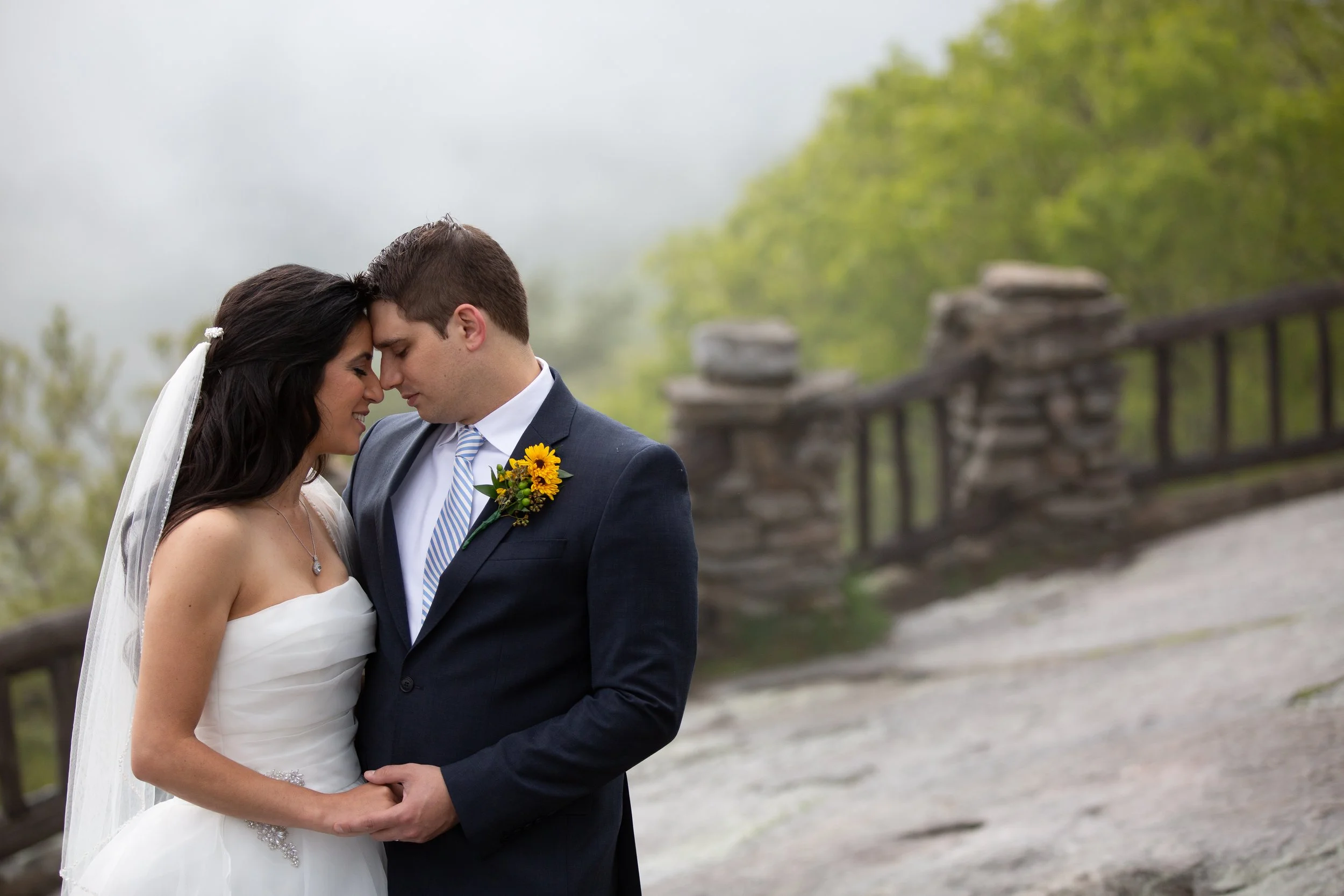 A bride and groom with foreheads touching, holding hands, standing outdoors near a wooden fence with green trees in the background, on a foggy day.