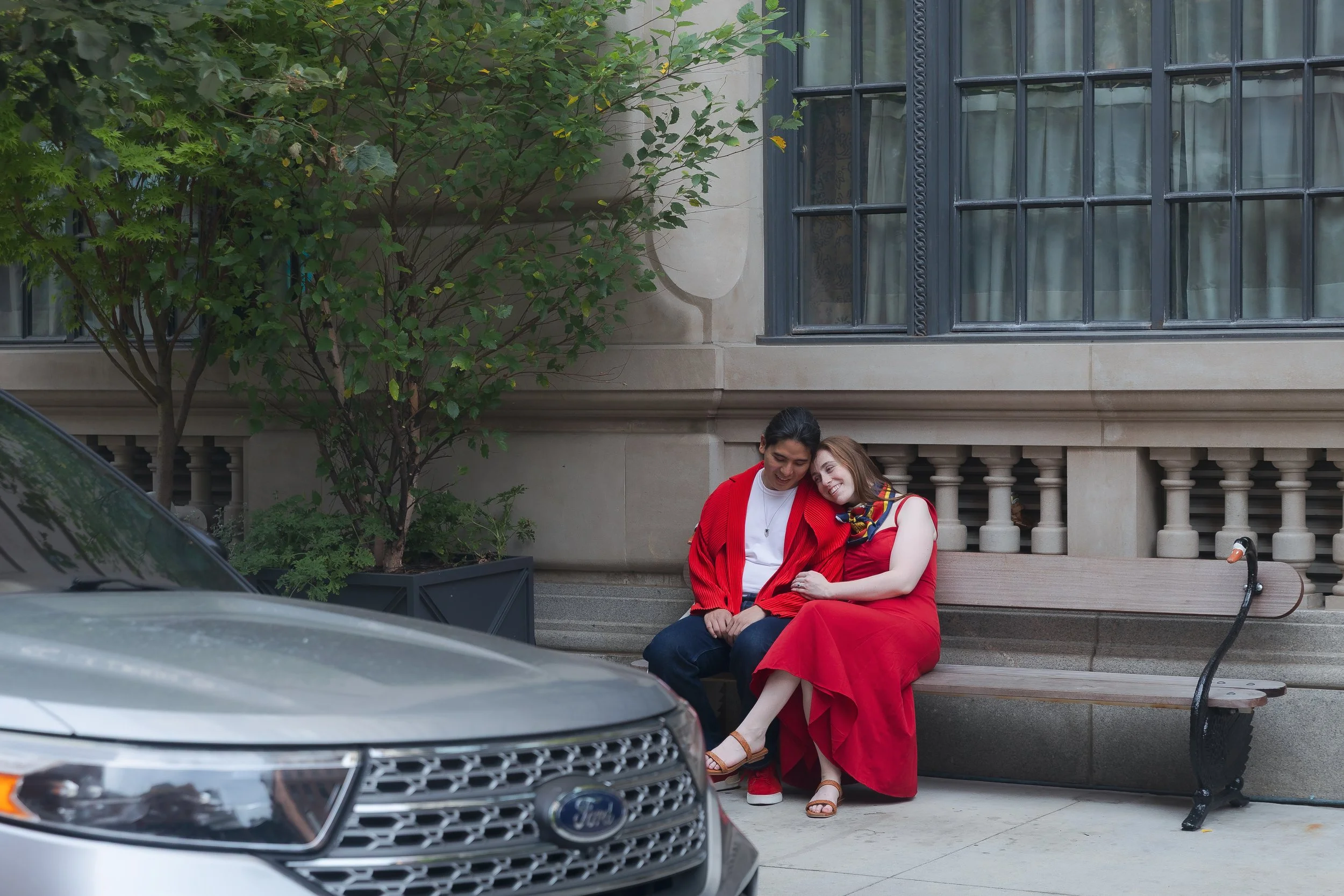 Two people sitting on a bench outside a building, smiling and leaning on each other. The person on the left is wearing a red jacket and white shirt, while the person on the right is dressed in red pants, a white top, and a colorful scarf. There is a 