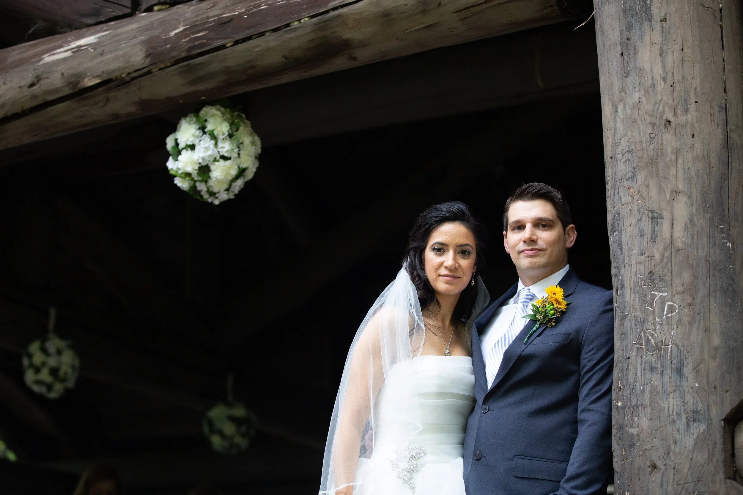 A bride and groom standing close together outdoors under a wooden structure, with a floral arrangement hanging above them. The bride is wearing a white wedding dress and veil, and the groom is wearing a dark suit with a striped tie and a boutonnière.