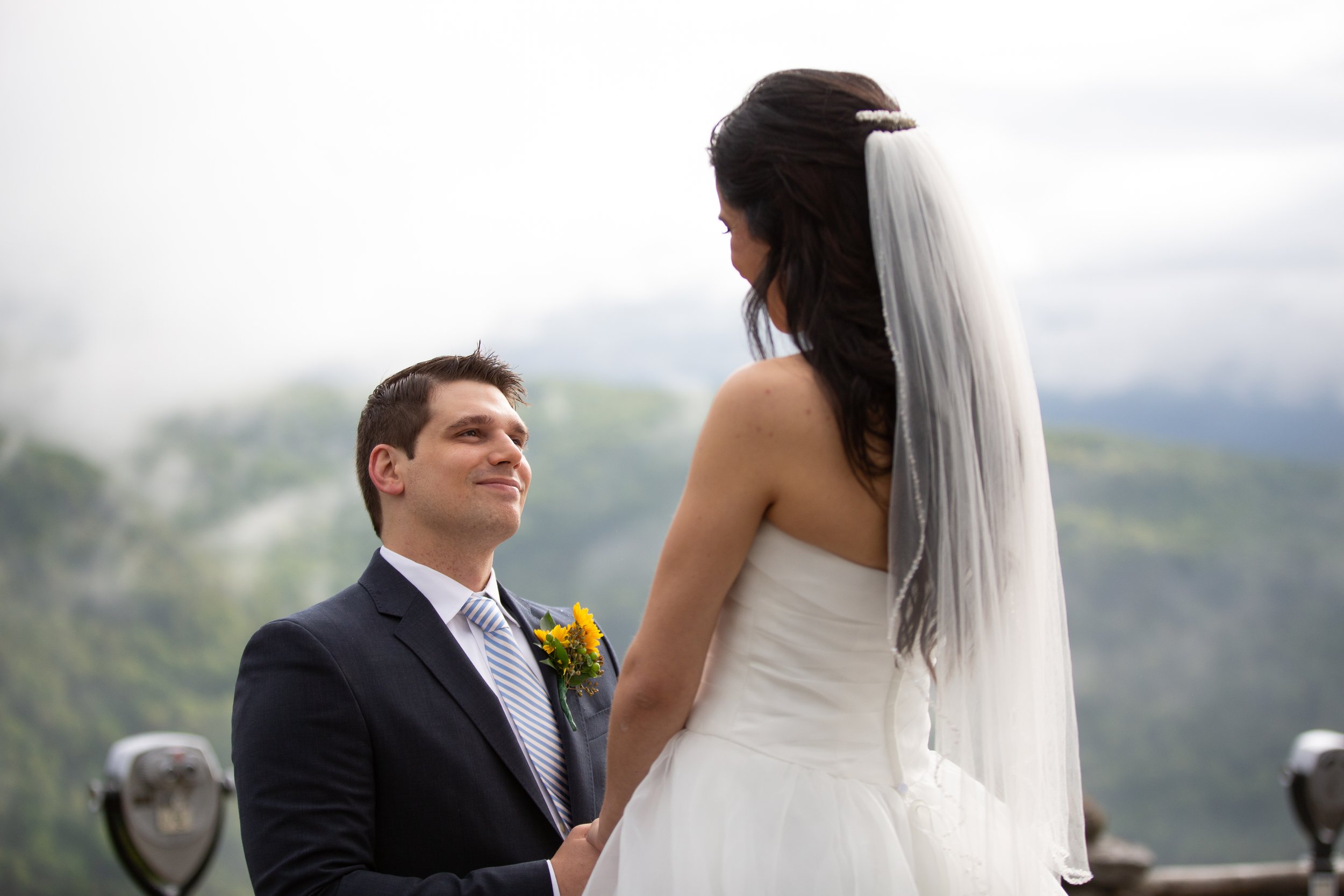 A bride and groom outdoors during their wedding ceremony. The groom in a black suit looks happily at the bride, who is wearing a white wedding dress and veil, overlooking a scenic landscape with cloudy sky.