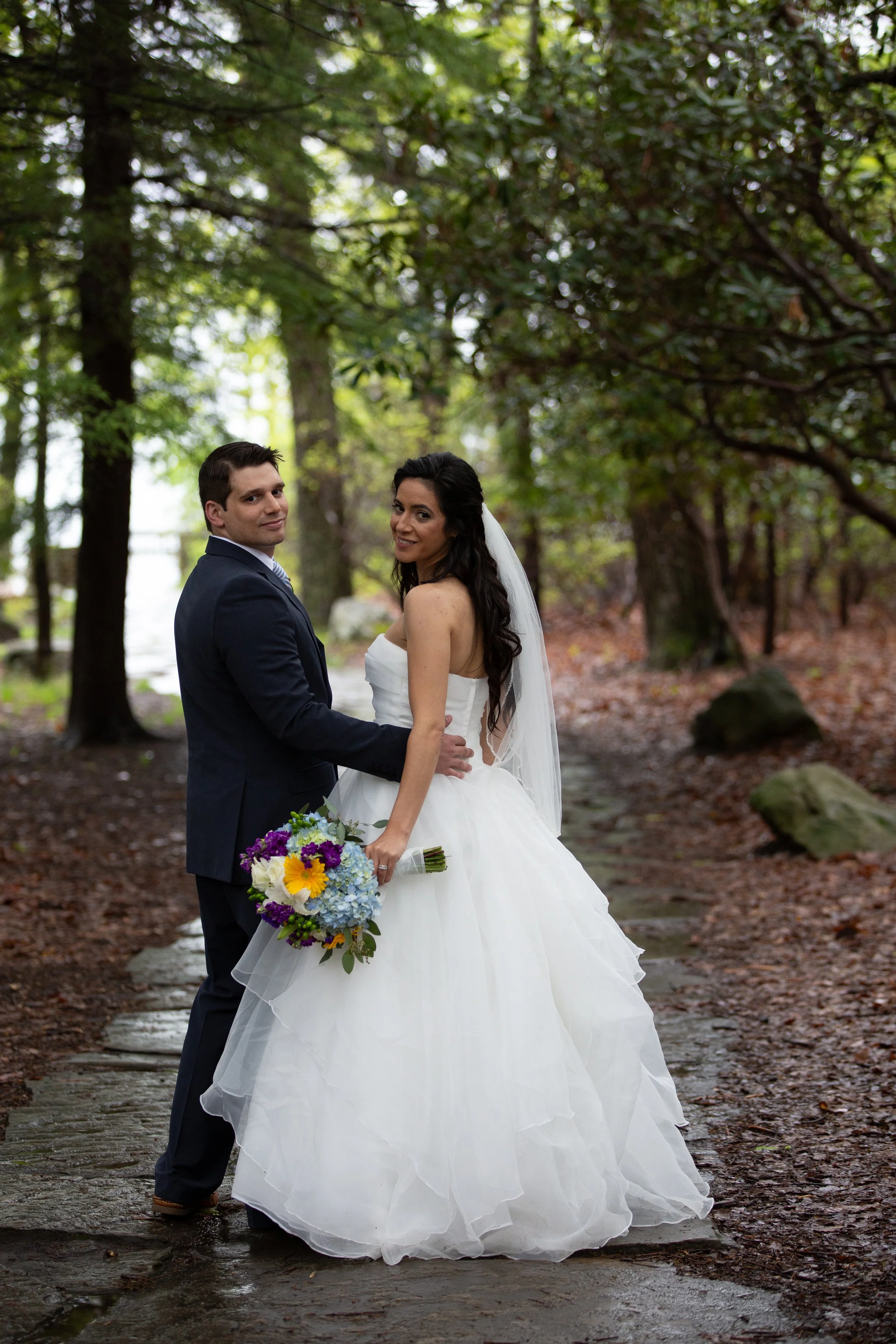 A bride and groom stand together holding hands in a wooded outdoor setting during their wedding, with the bride holding a colorful bouquet and wearing a white wedding gown. The groom is dressed in a dark suit. The background features trees and greene
