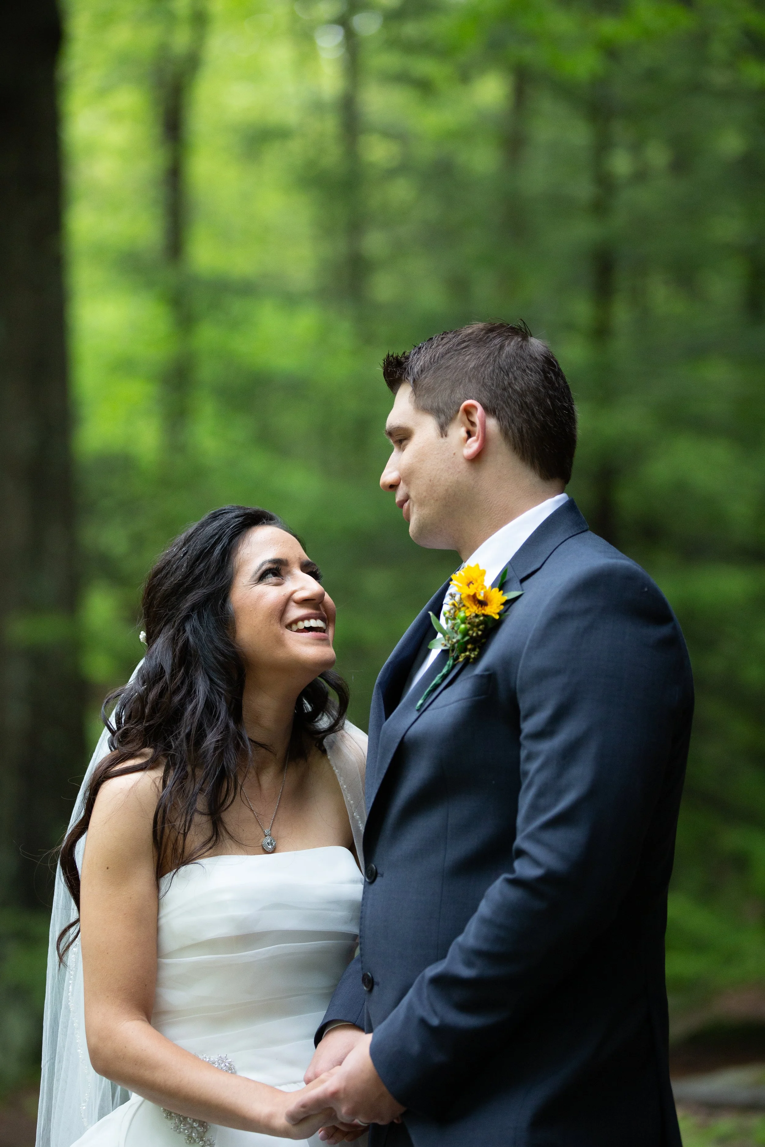 A bride and groom standing outdoors in a forest, looking at each other and smiling, holding hands.