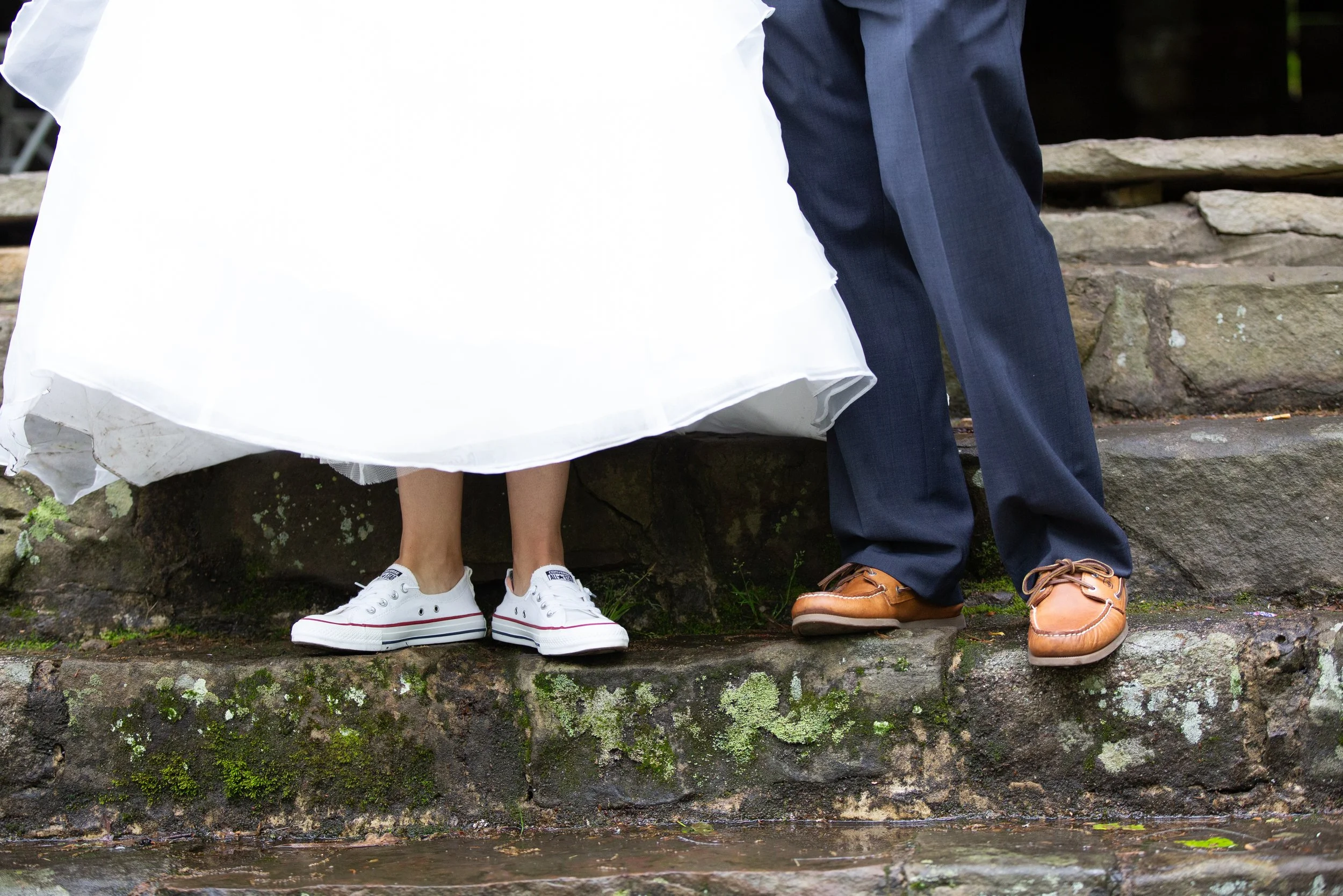 Lower half of two people sitting on a stone wall, one wearing white sneakers and a white dress, the other wearing brown loafers and navy blue pants.