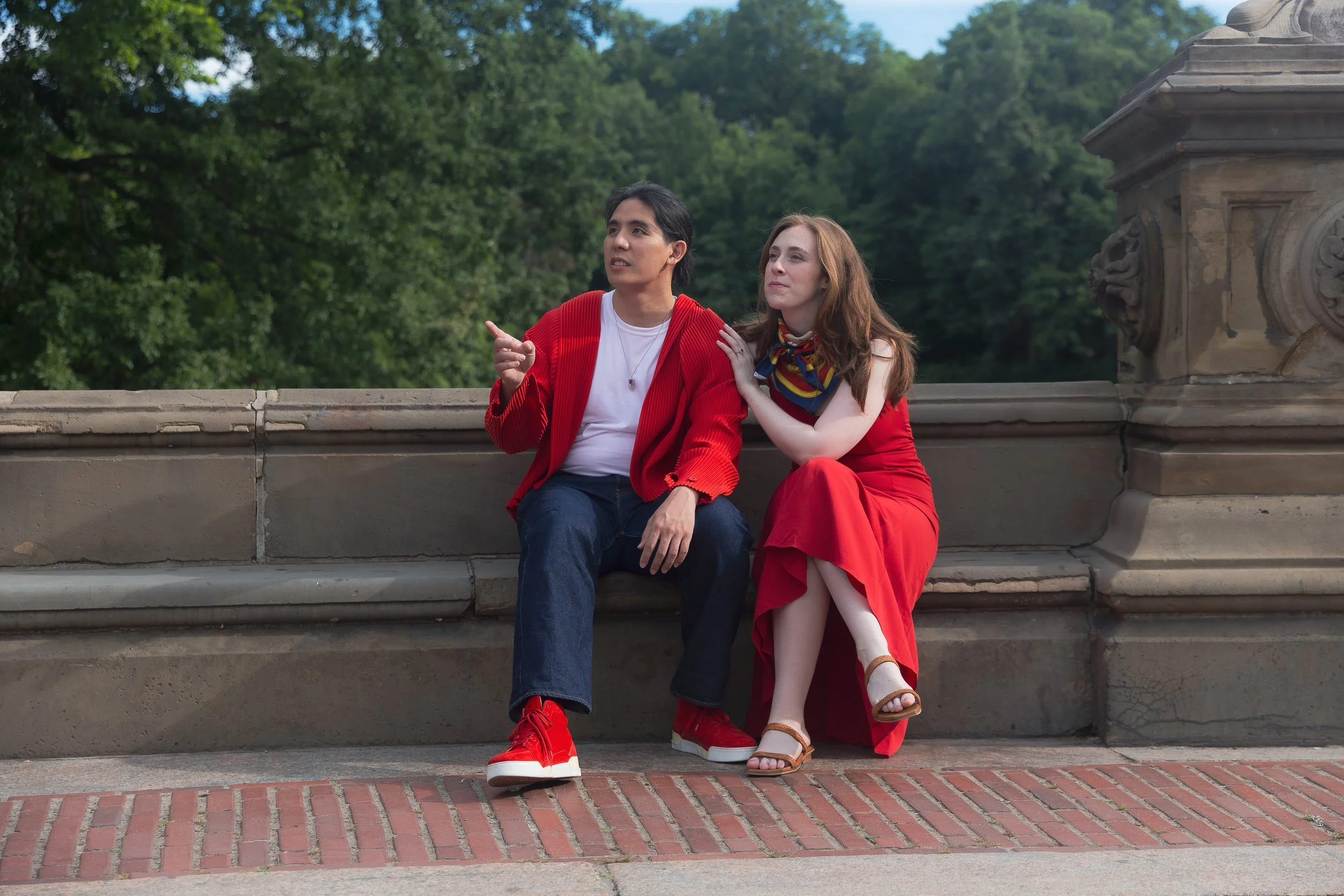 Two people sit on a stone bench outdoors, one appears to be explaining or pointing, both wearing red clothing, with a lush green background.