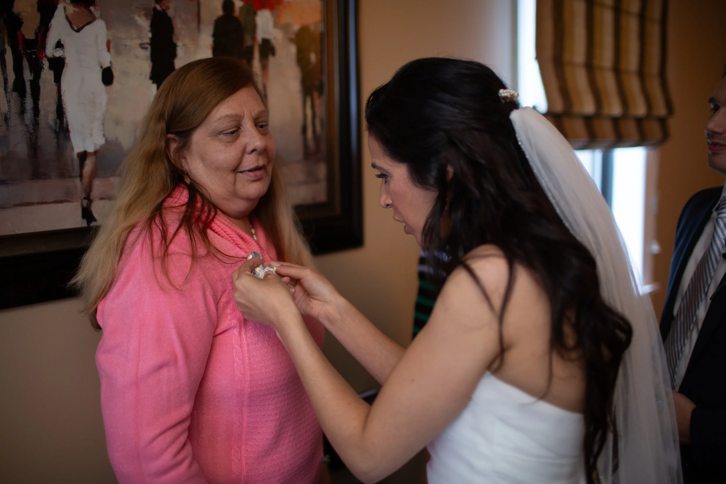 A woman in a white dress, likely a bride, is pinning a boutonniere on a woman in a pink sweater, who appears to be the mother of the bride. Two other people are partially visible in the background.