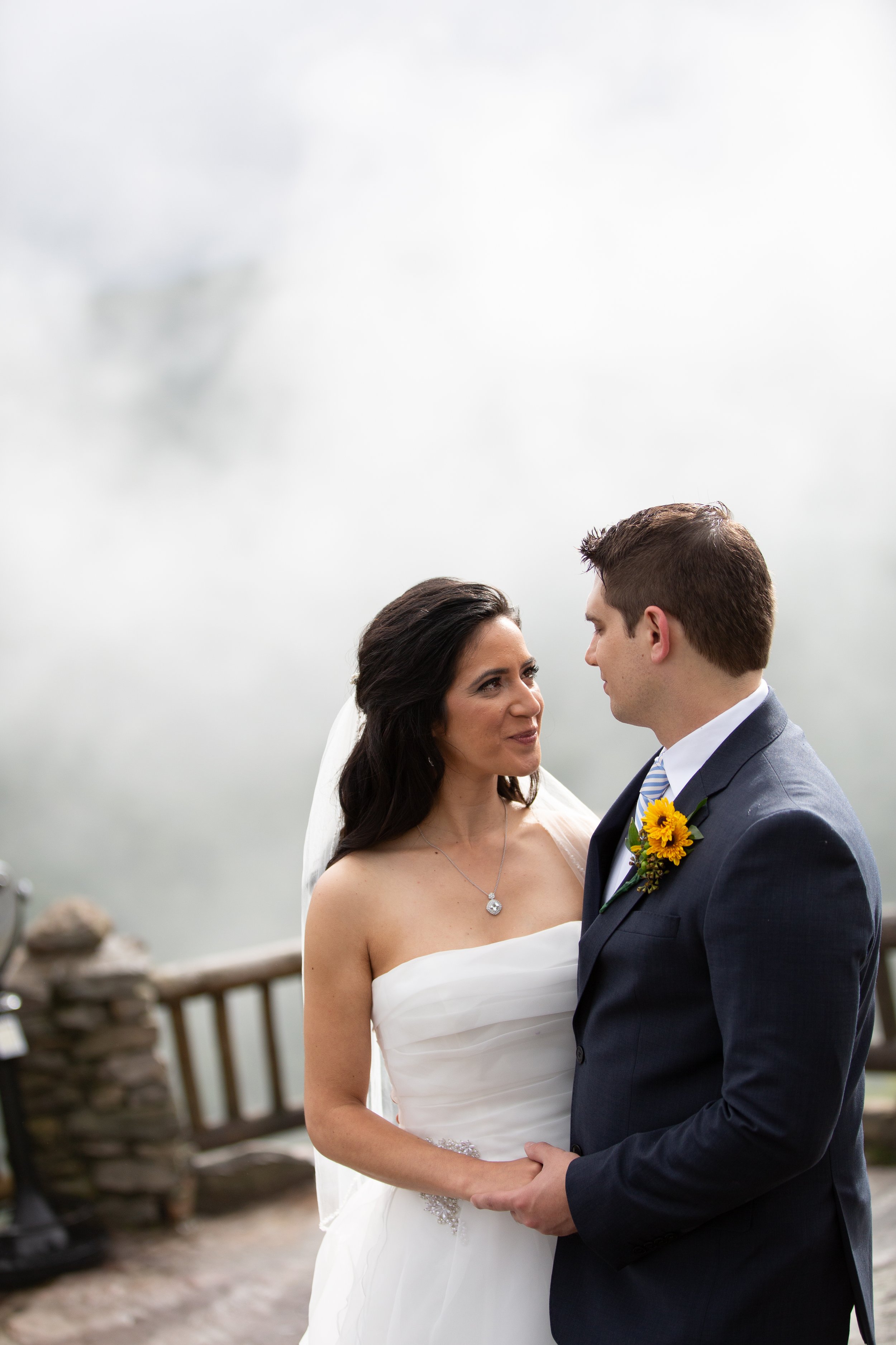 A bride and groom standing close together outdoors during their wedding ceremony, holding hands and gazing at each other, with a cloudy sky above.