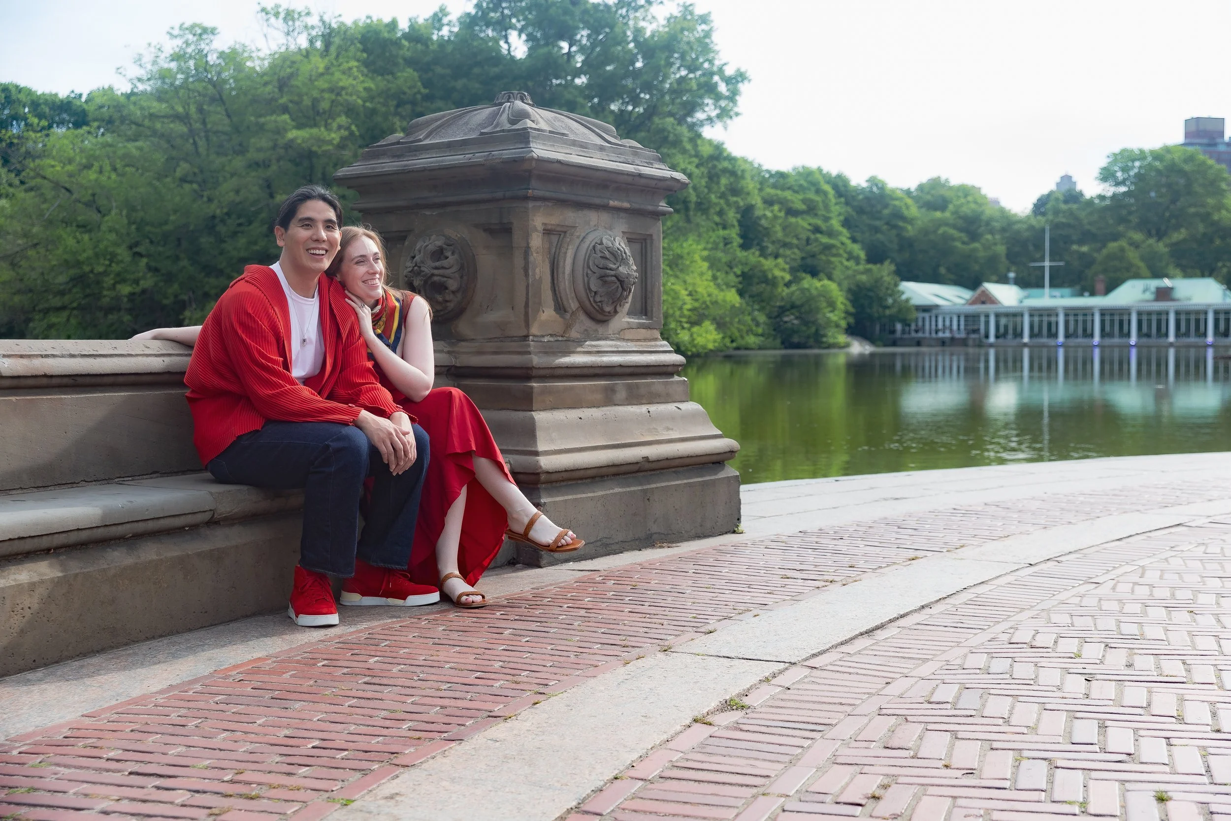 A young man and woman sitting on a brick and stone ledge by a river, smiling and leaning close together. The man wears a red jacket, white shirt, and dark pants, while the woman wears a patterned top and a long red skirt. Behind them is a stone pilla