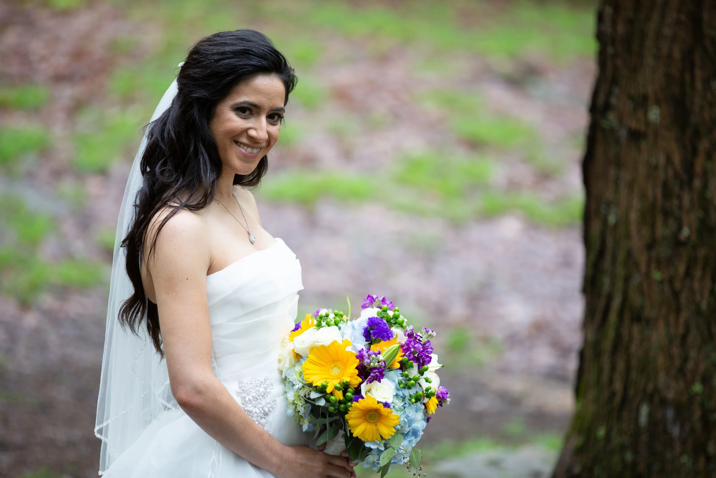 A bride with long dark hair wearing a white strapless wedding dress and veil, holding a bouquet of colorful flowers, standing outdoors near a tree in a forested area.