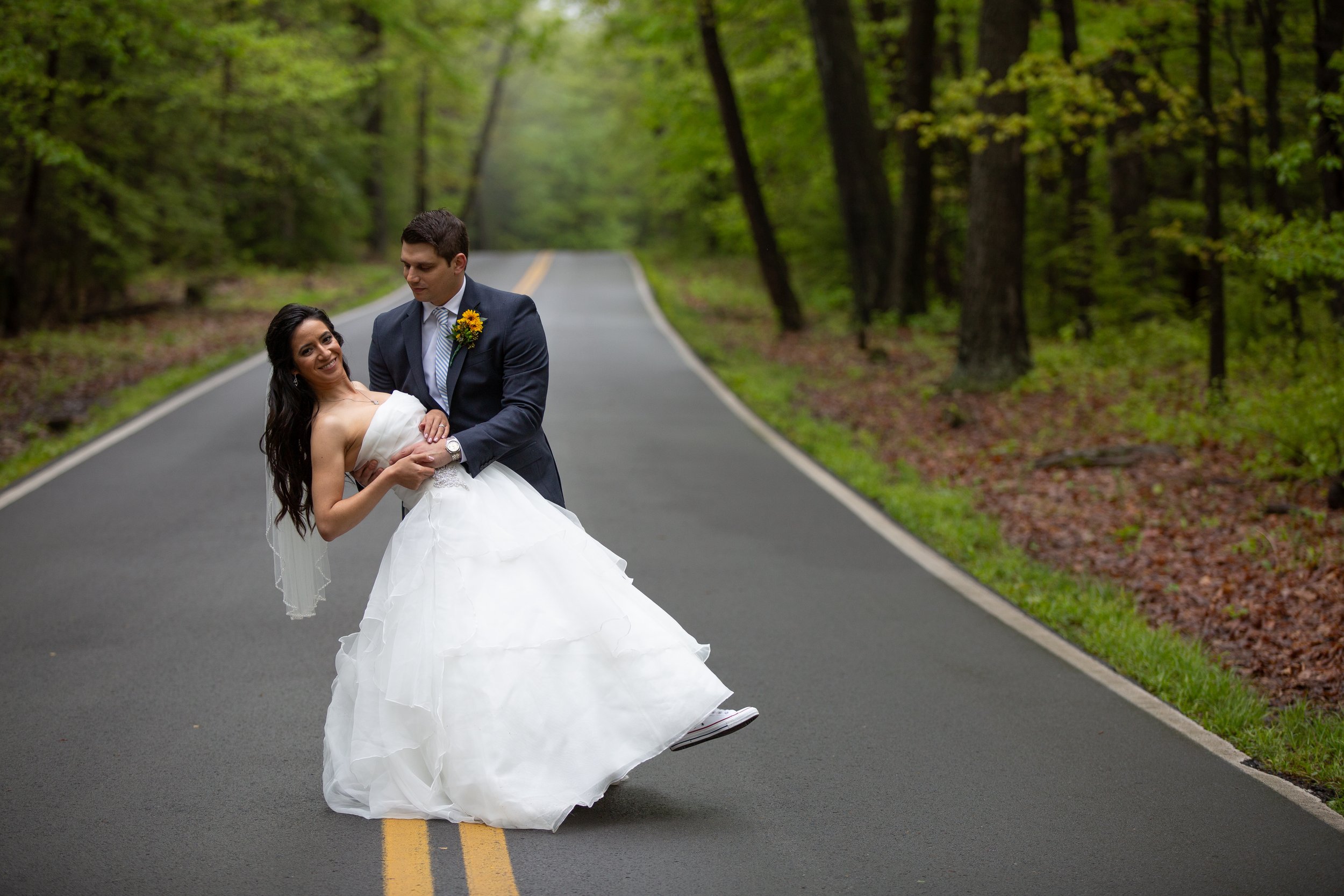 A bride and groom dancing in the middle of a forest road surrounded by green trees.