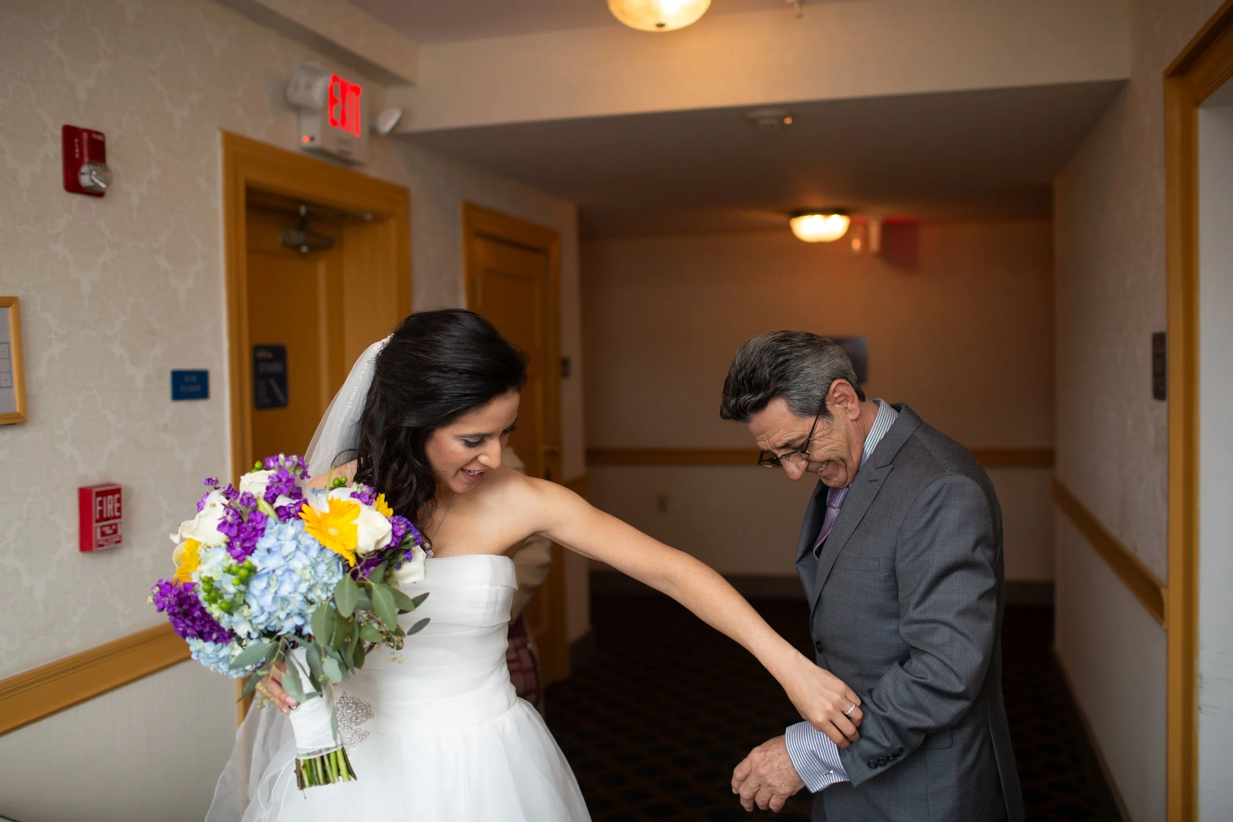 A bride in a white wedding dress holding a bouquet of purple, yellow, and white flowers, while an older man, likely her father, is bending down and smiling.