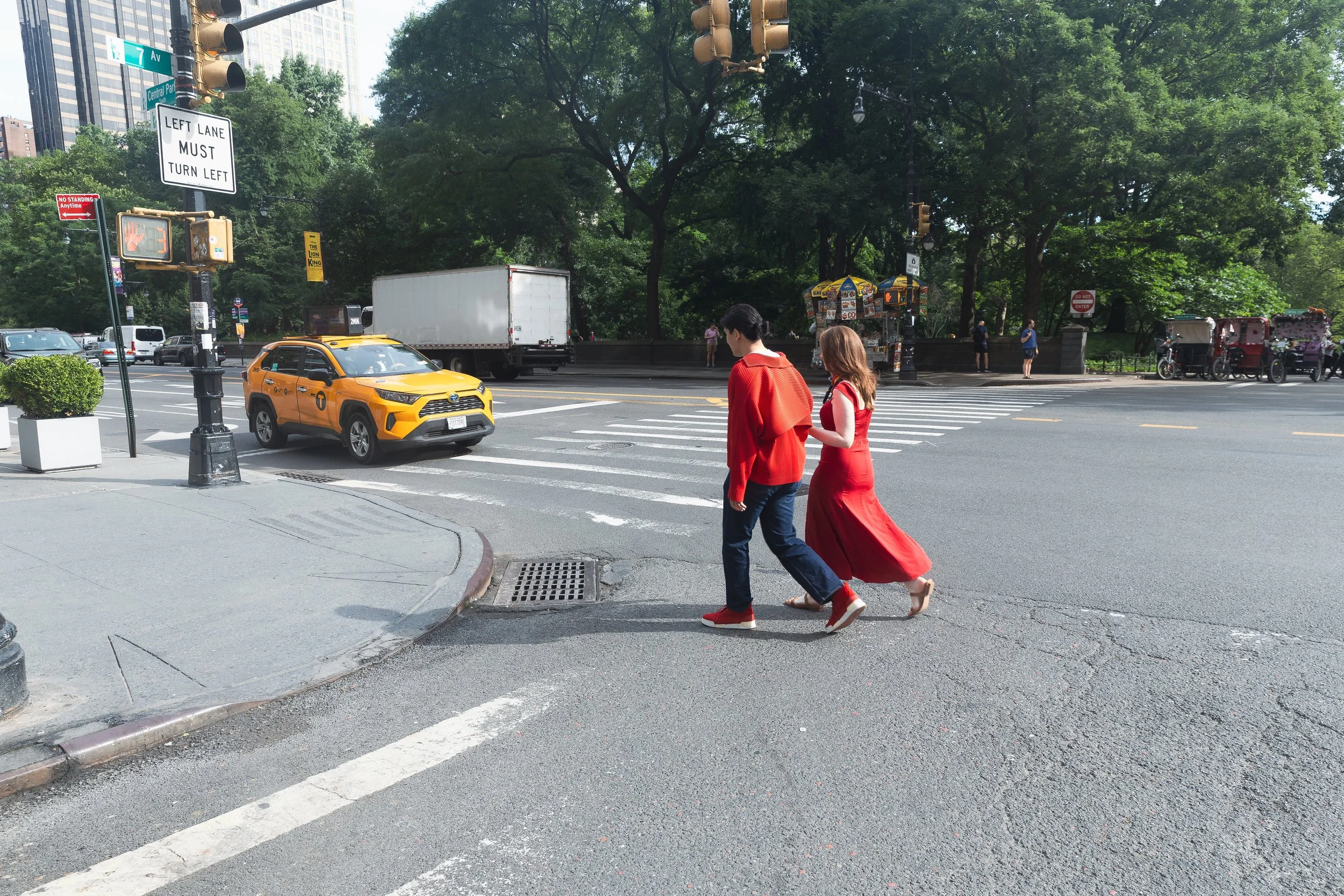 Two people are crossing a city street at a crosswalk, one in a red dress and the other in a red jacket and jeans, with cars and trucks passing by and trees in the background.