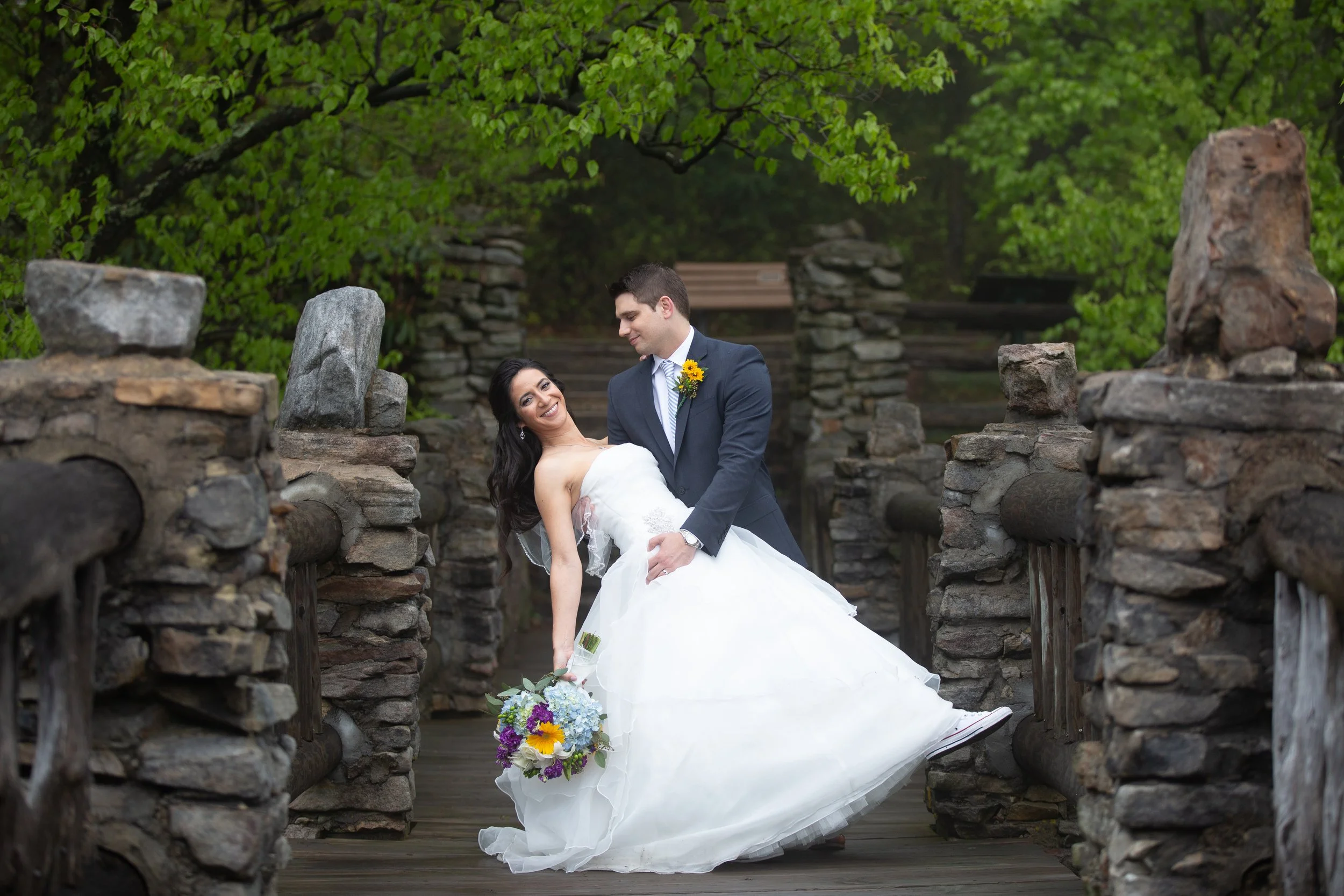 A bride and groom on a stone bridge surrounded by lush green trees, with the bride smiling and leaning back as the groom looks at her. The bride holds a bouquet of colorful flowers.