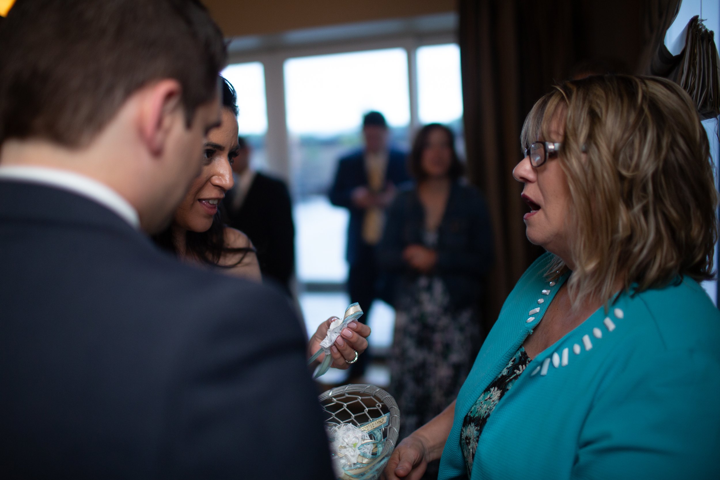 A woman with glasses receiving a gift or keepsake from a person in a suit at a social gathering, with other people in the background near a window.