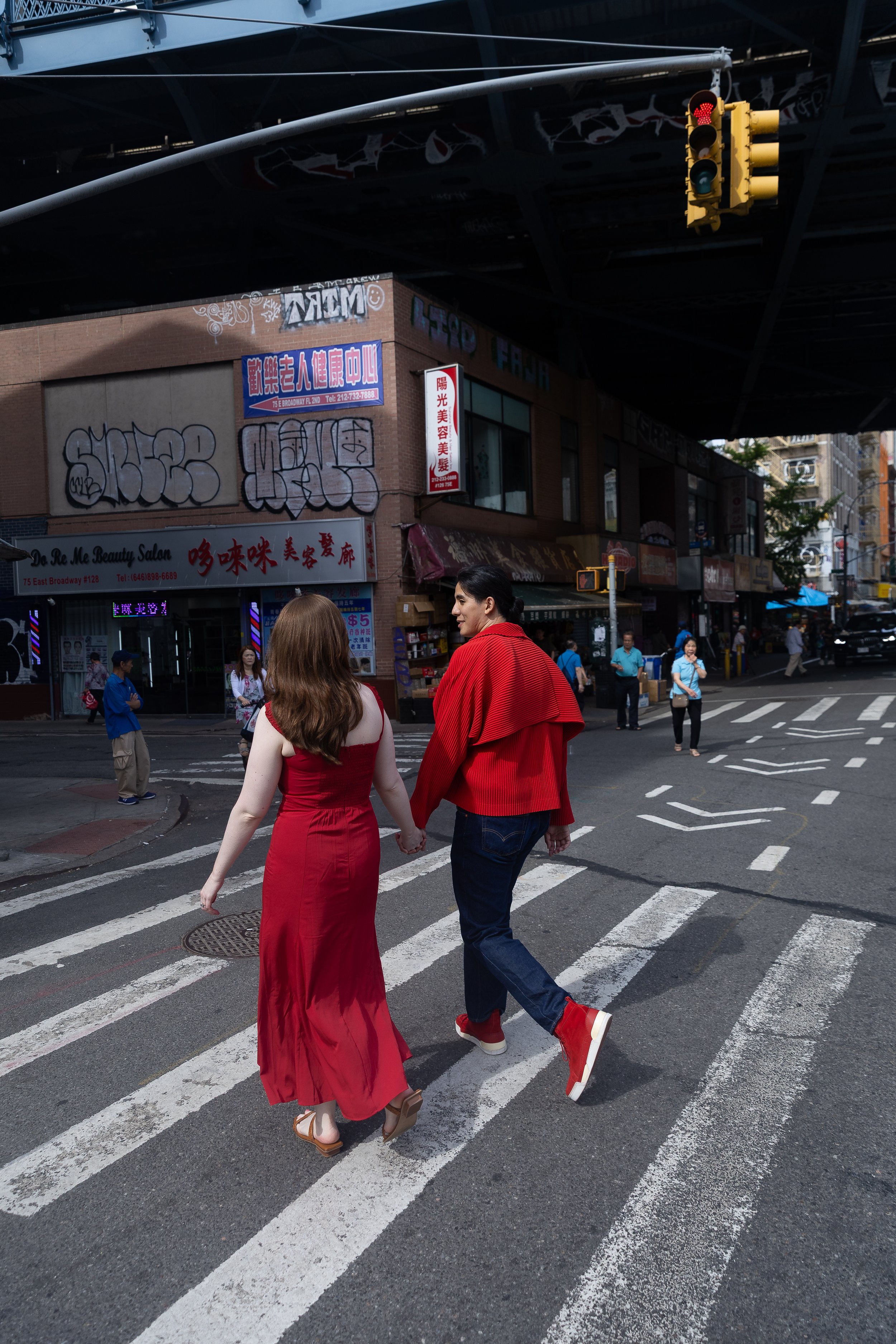 A woman in a red dress and a man in a red jacket walk hand in hand across a crosswalk in an urban area with multiple pedestrians and buildings, under a bridge with traffic lights.