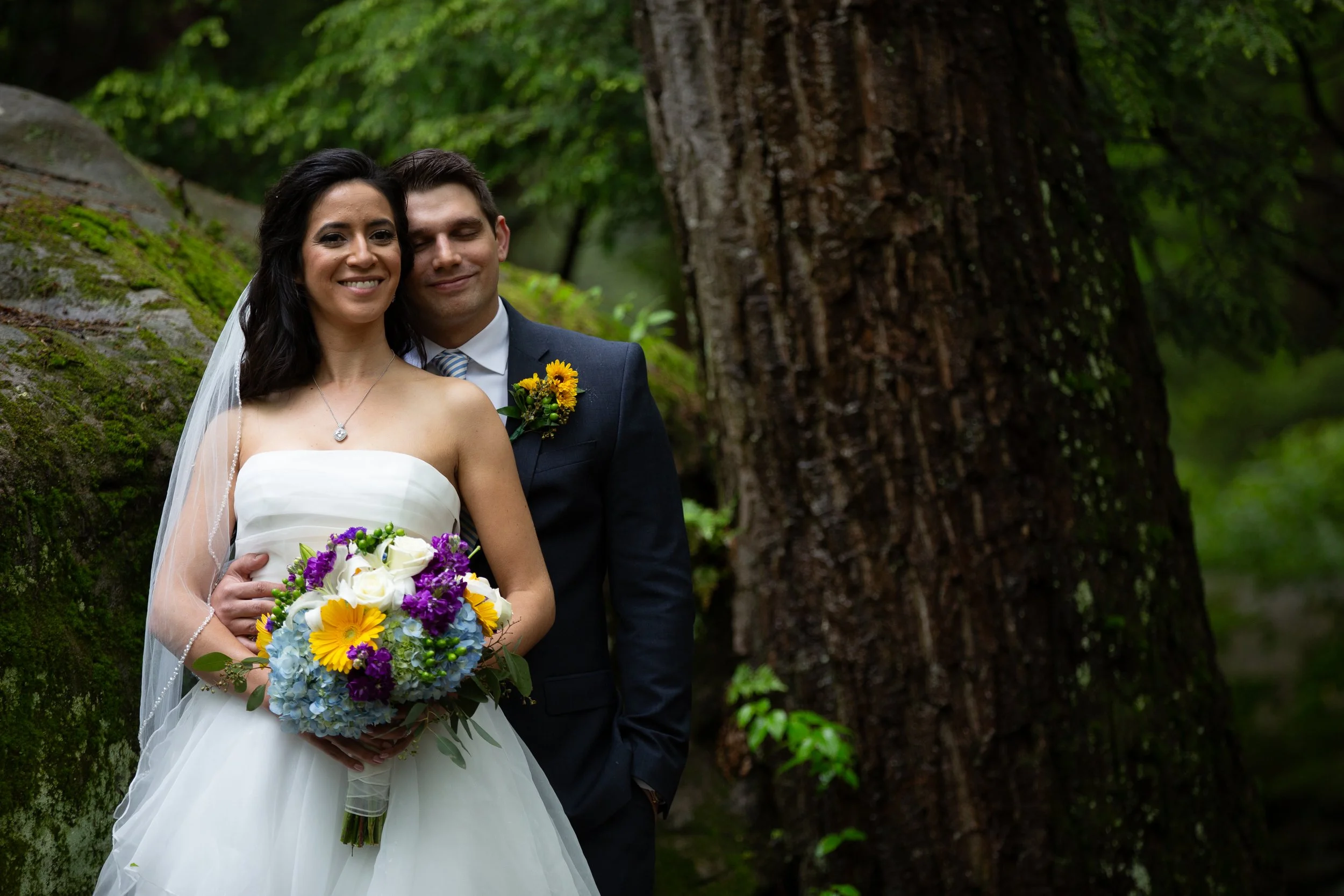 A newlywed couple standing outdoors in a forest, smiling. The bride is holding a colorful bouquet of flowers, and the groom is standing close behind her with his eyes closed, wearing a dark suit with a boutonniere.