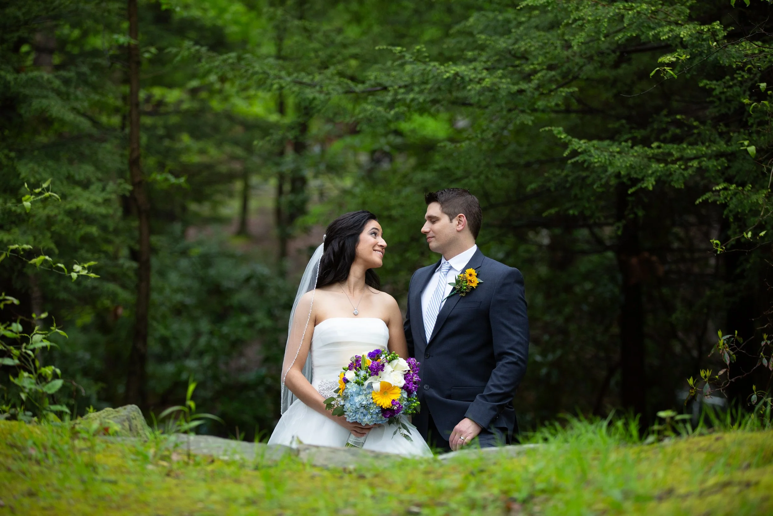 A bride and groom standing outdoors in a lush green forest, smiling and gazing at each other, with the bride holding a colorful bouquet.