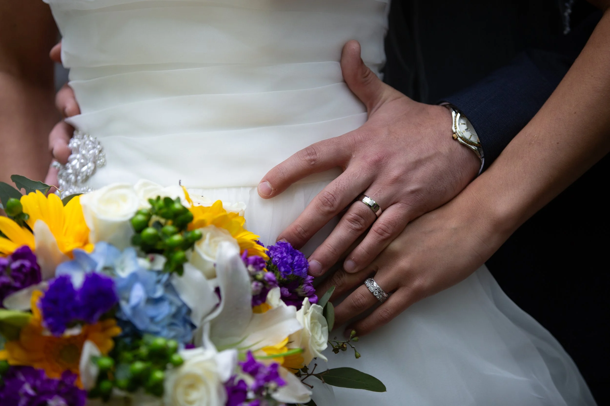 Close-up of a newlywed couple's hands on their wedding day. The bride's hand, adorned with rings, rests on top of the groom's hand, which is also wearing a ring. The groom is wearing a wristwatch. The couple's wedding dress and suit are visible, alon