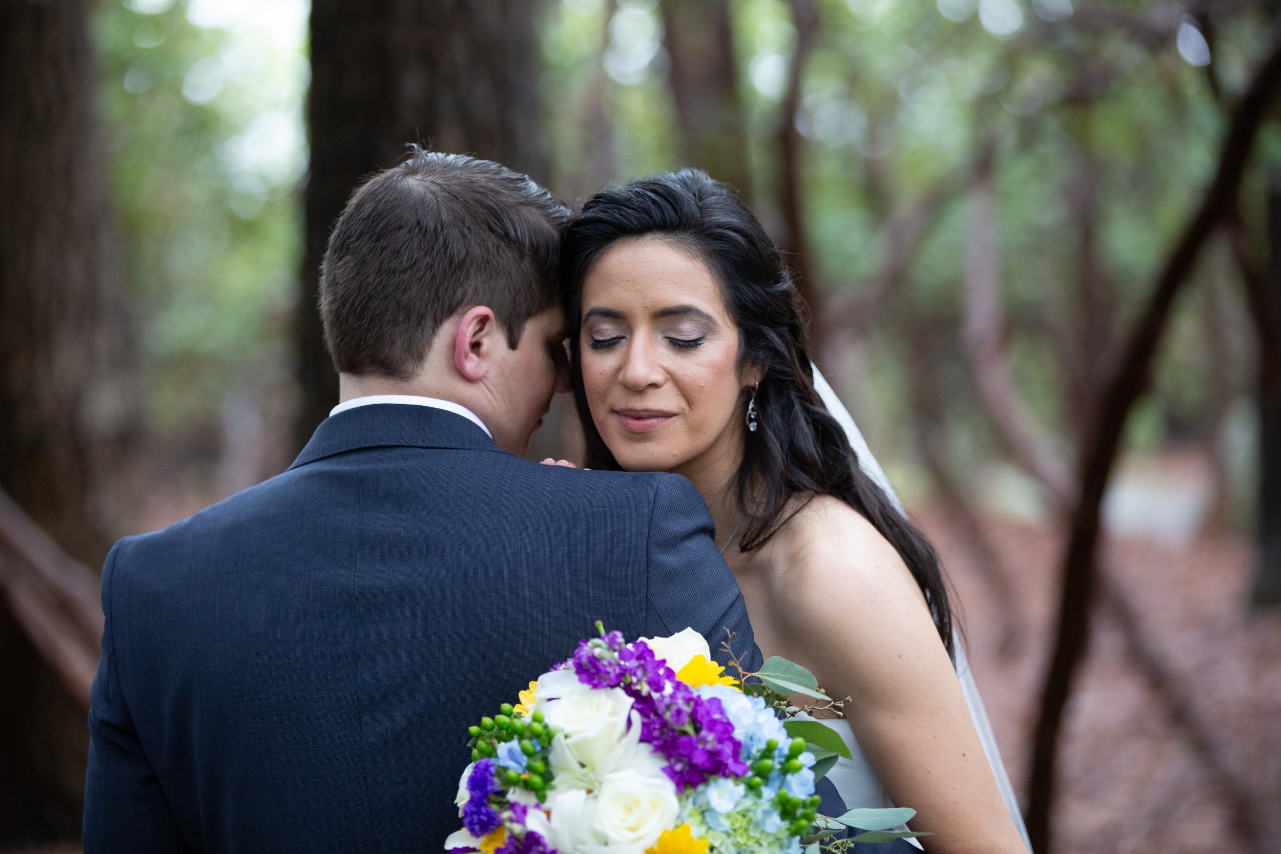 A bride and groom share a tender moment outdoors, with the groom whispering to the bride and holding a colorful bouquet of flowers, surrounded by a blurred wooded background.