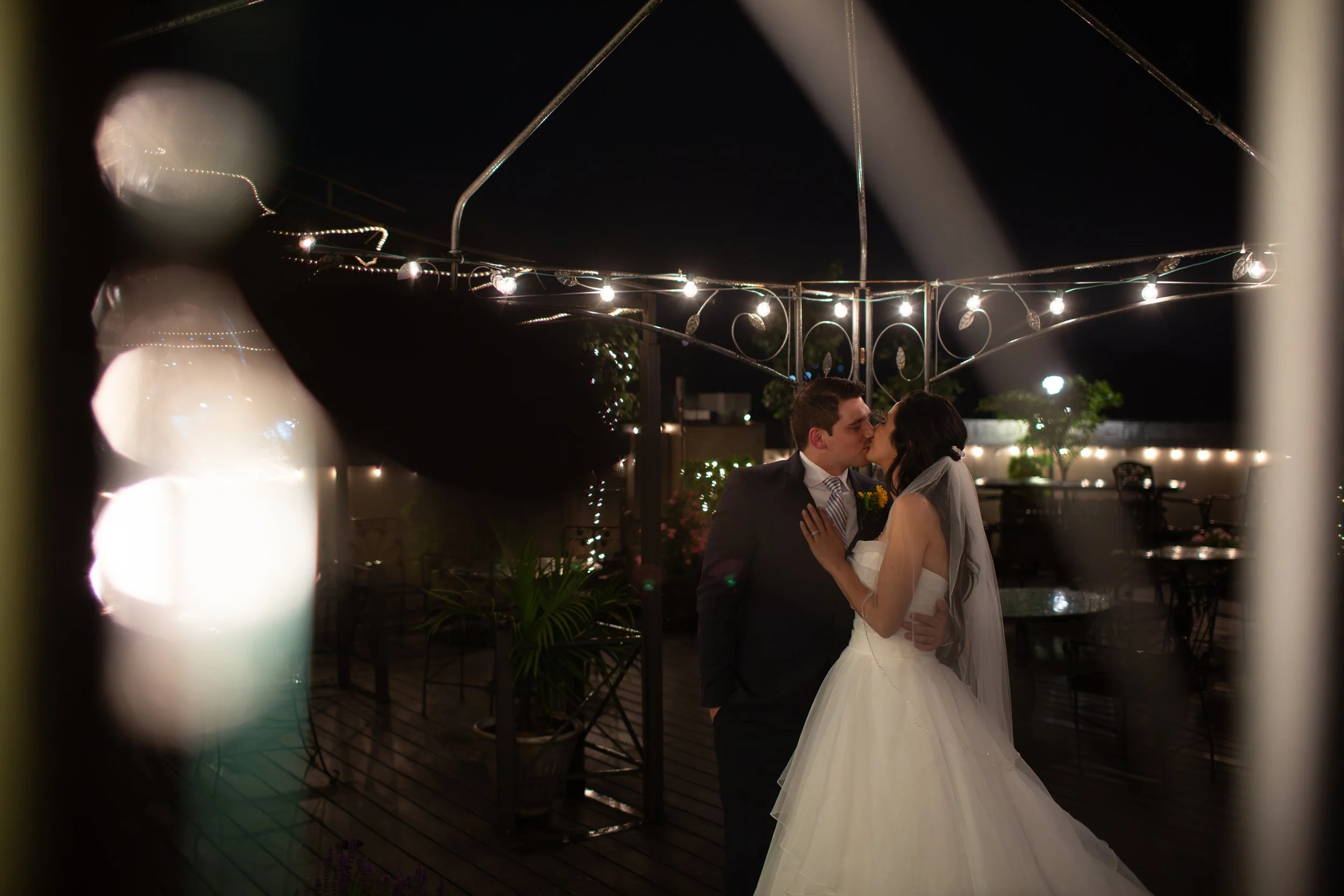 A bride and groom kissing at their wedding at night with string lights overhead, viewed through a blurry foreground.